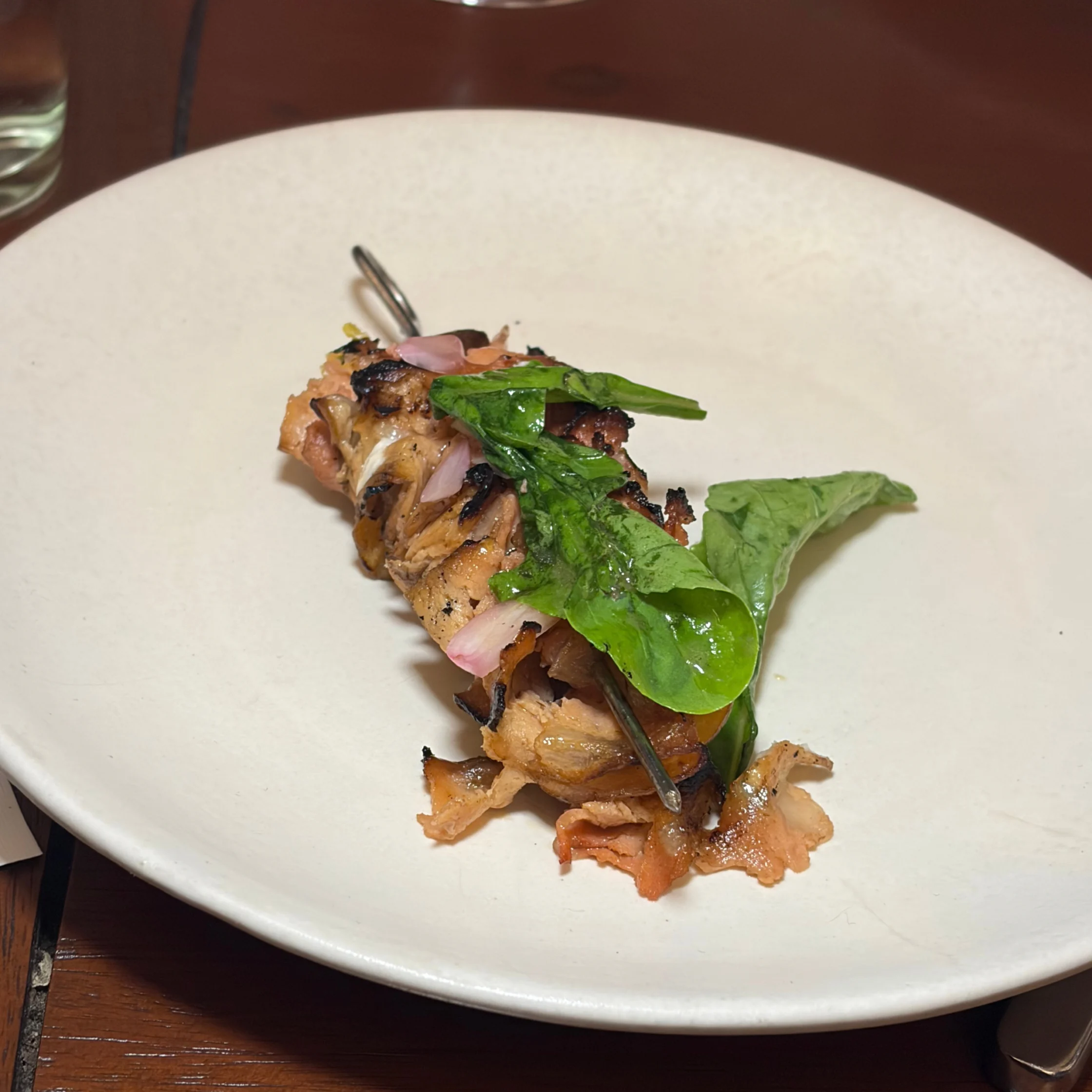 Grilled chicken skewers topped with fresh green leaves and sliced shallots sit on a large white plate, creating a simple, appetizing restaurant presentation.