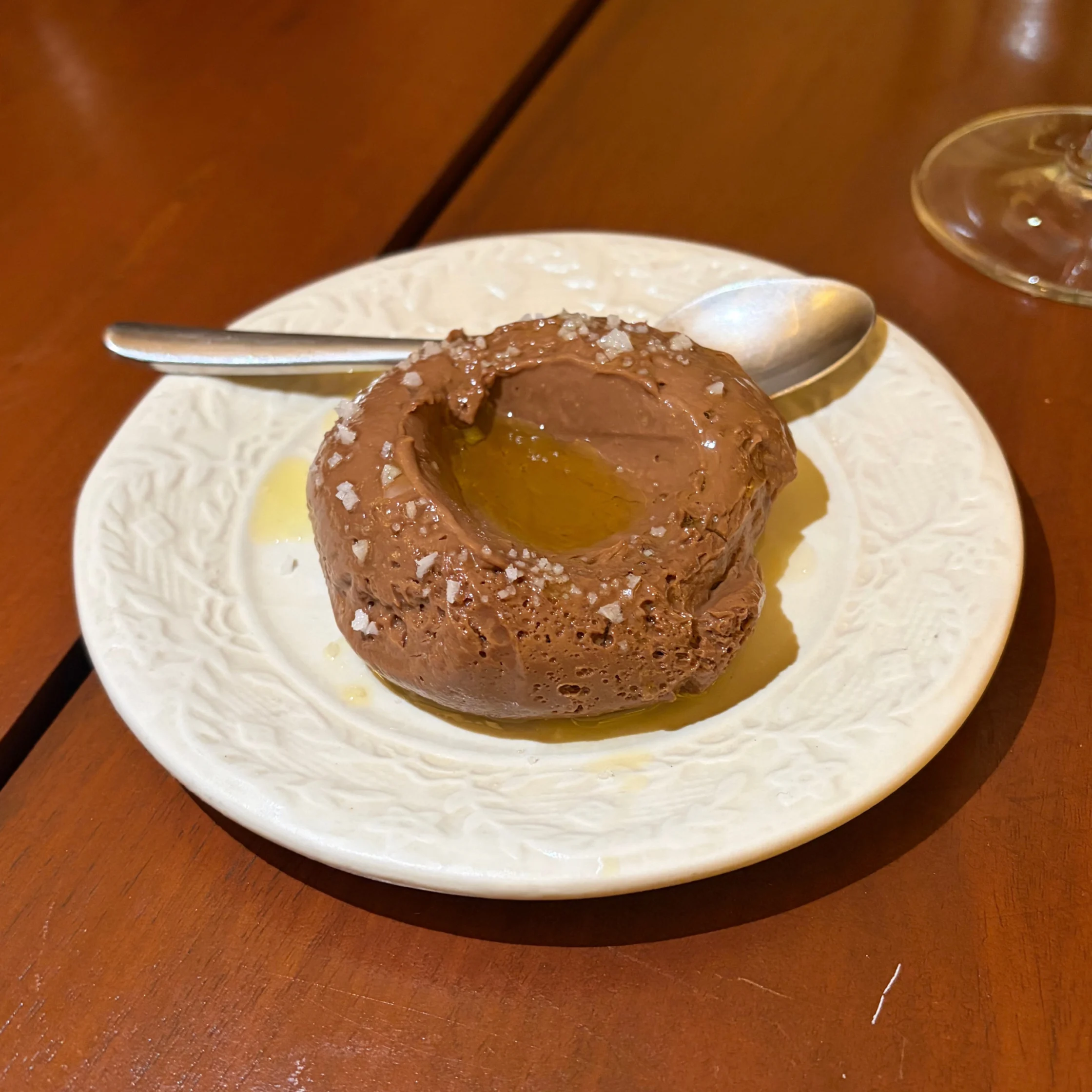 A rich chocolate lava cake dusted with coarse salt sits on a white plate, its warm golden center exposed; a spoon rests behind it on a wooden table.
