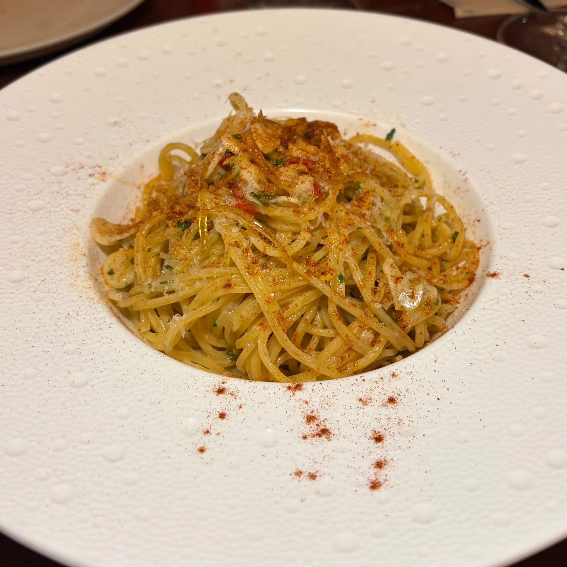 A gourmet plate of golden spaghetti with herbs and grated cheese, centered in a wide white dish dusted with red seasoning; elegant, appetizing presentation.