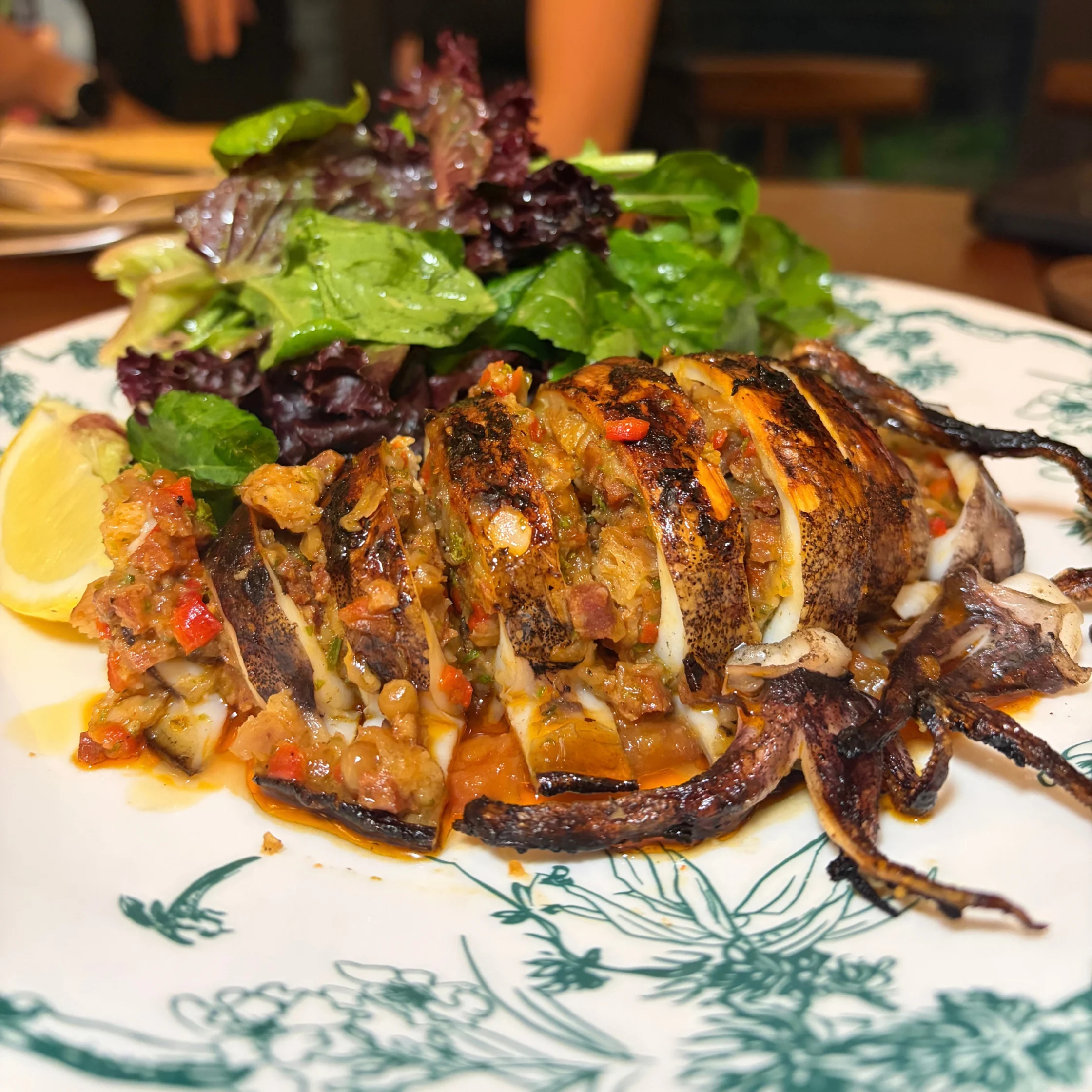 A richly browned, sliced grilled squid stuffed with vegetables and herbs, served with leafy greens and lemon on a patterned plate; warm, appetizing restaurant scene.