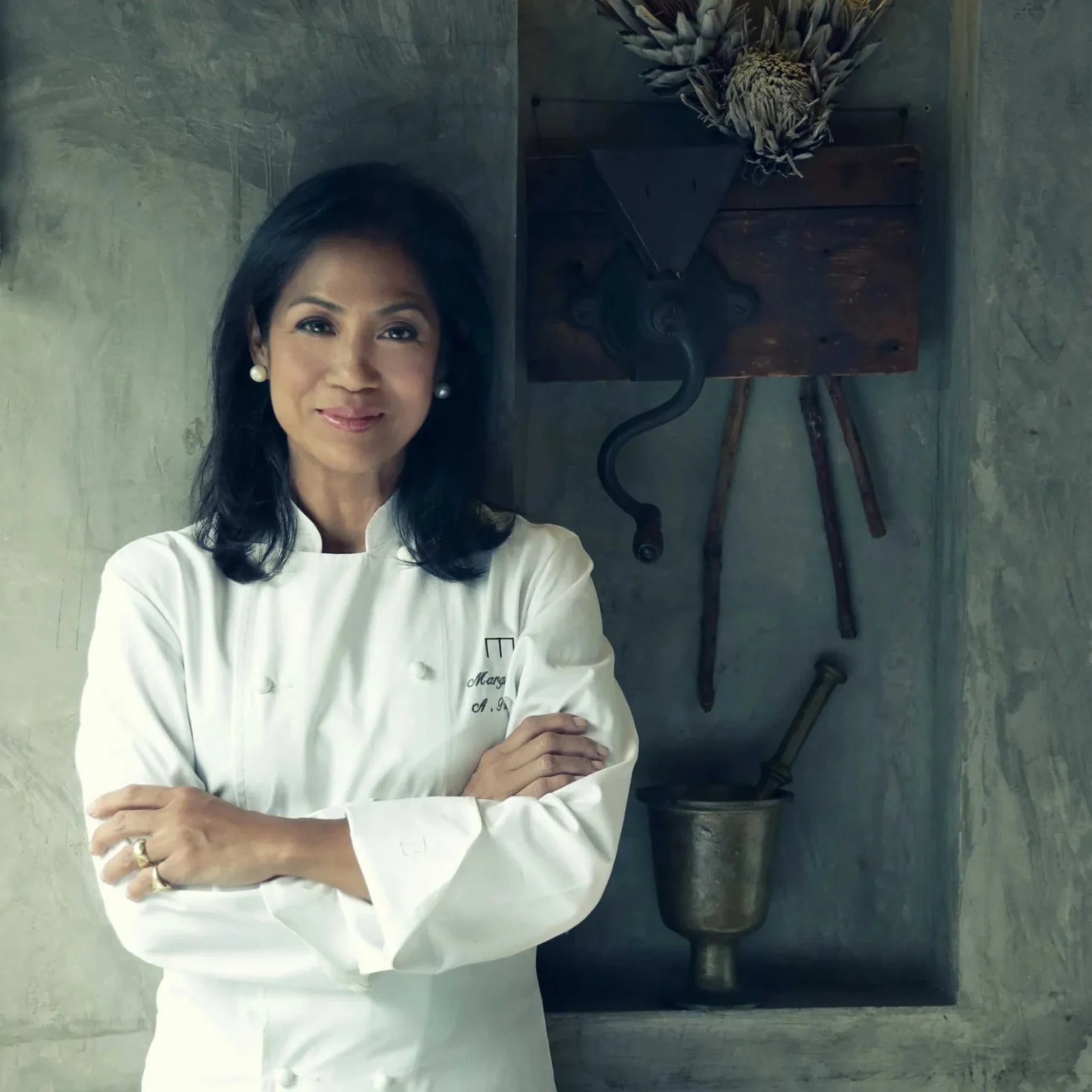 A woman chef in a white coat stands with arms crossed in a rustic kitchen, smiling calmly beside an old stove and hanging herbs.