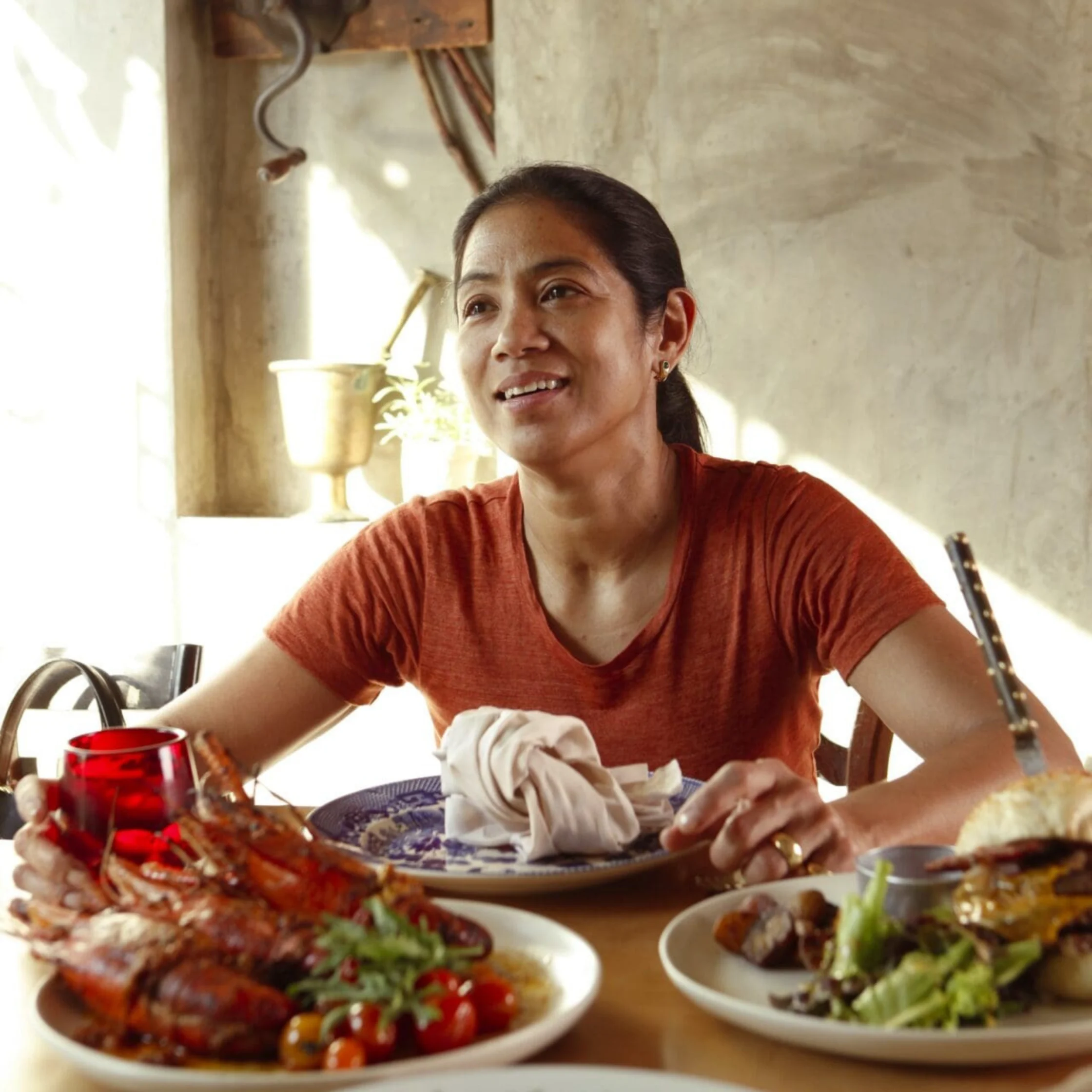 A smiling woman in a rust-red shirt serves food at a table laden with roasted seafood, salad, and a red drink in a warmly lit home.