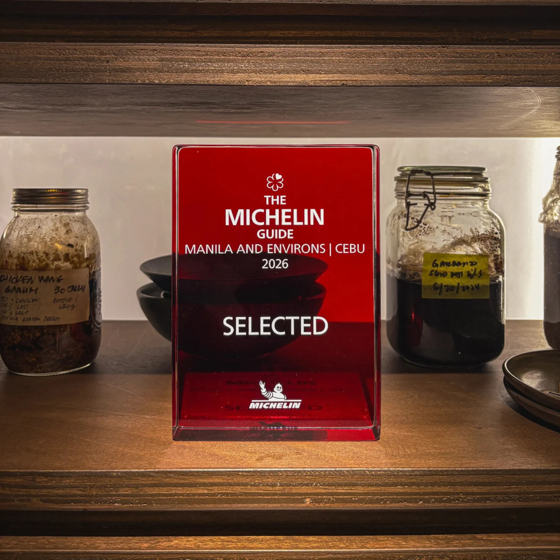 Red Michelin Guide “Selected 2026” award stands on a wooden shelf between glass jars and dark bowls, creating a warm, rustic restaurant display.