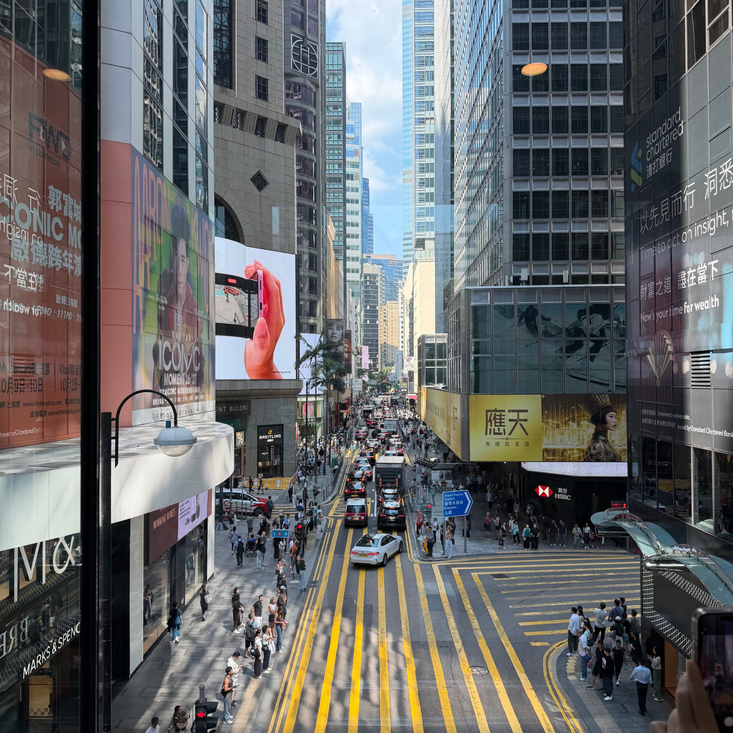 Elevated view of a busy Hong Kong street lined with towering glass buildings, colorful billboards, taxis, buses, and crowds crossing yellow road markings; energetic city bustle.