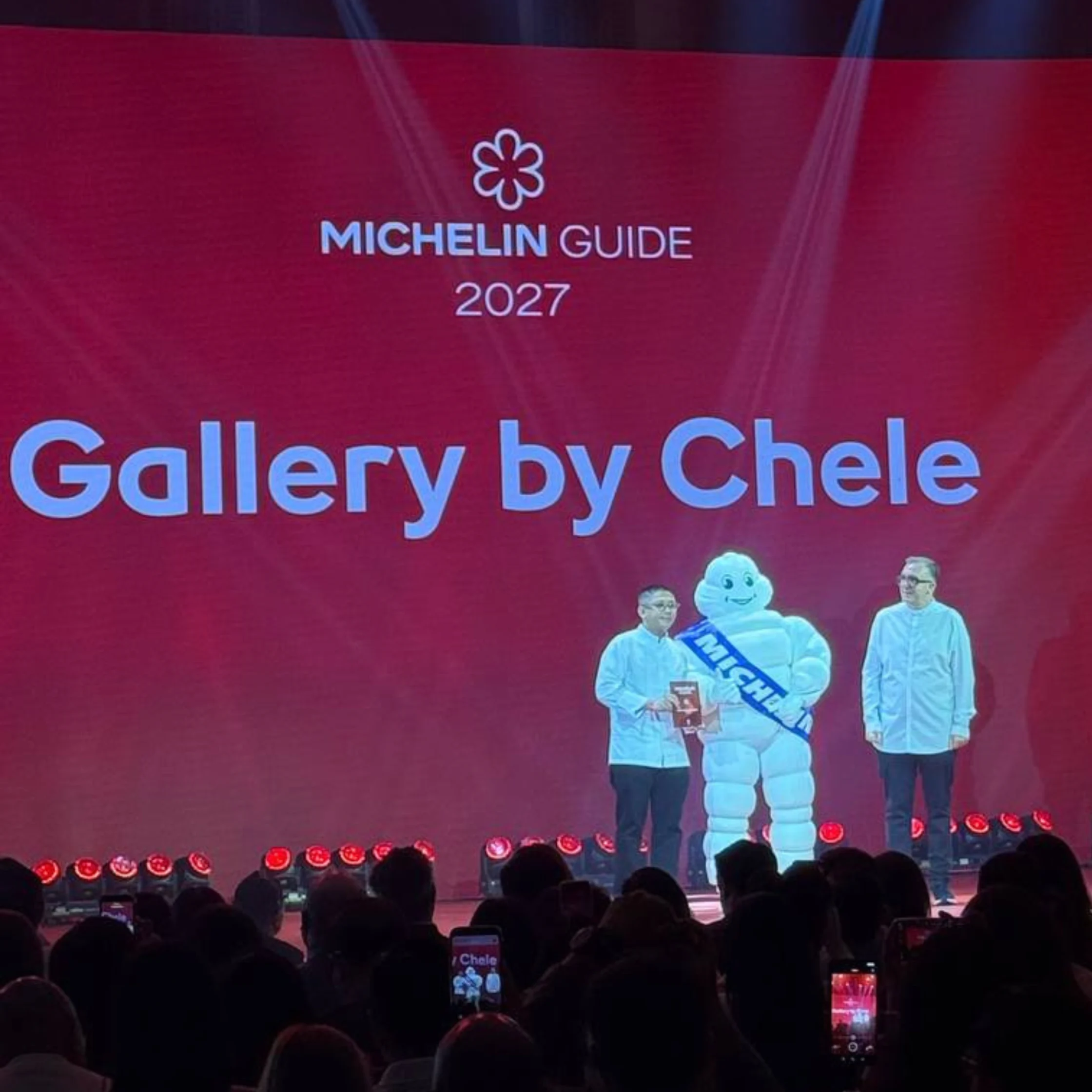 Michelin Guide 2027 stage announcement for “Gallery by Chele,” with two men and the Michelin mascot before a red backdrop and audience. The mood is celebratory.