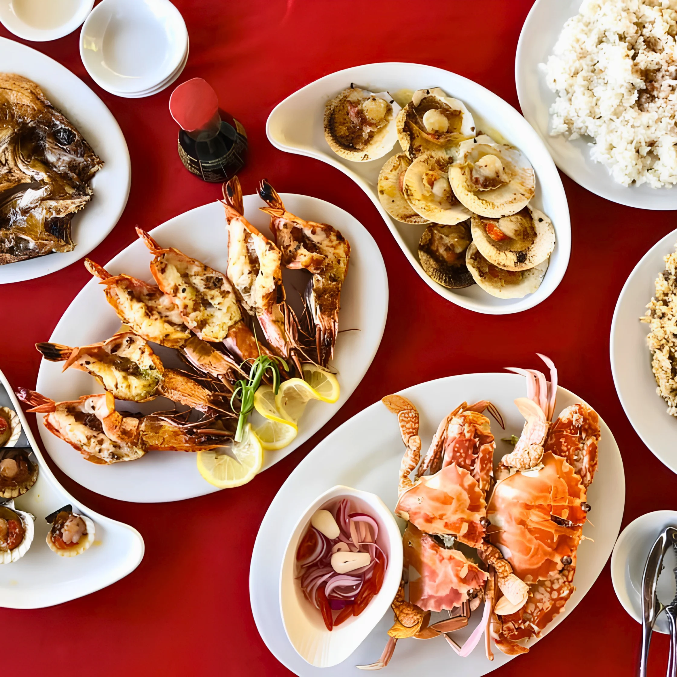 Overhead feast of grilled prawns, orange crab, scallops, and fish on white platters over a vivid red tablecloth, creating a festive seafood spread.