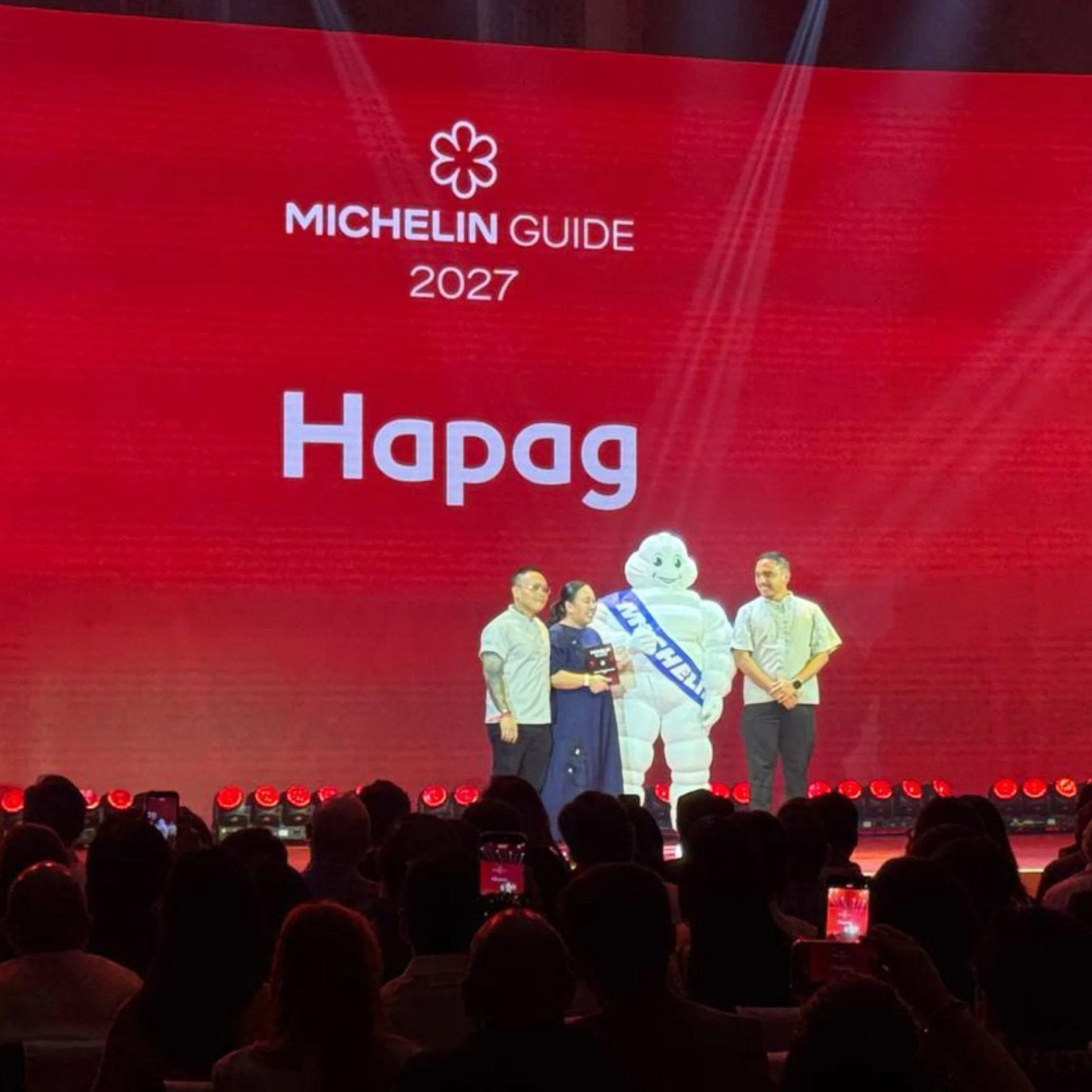 On a red Michelin Guide 2027 stage, four people stand beside the Michelin Man beneath the word “Hapag,” as a silhouetted audience watches.