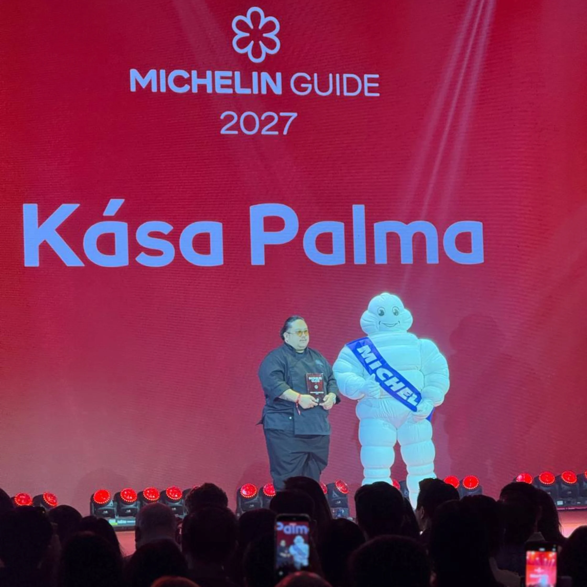 Michelin Guide 2027 stage display names Kása Palma, who stands beside the Michelin Man before a crowd; bright red backdrop and blue lettering create a celebratory tone.