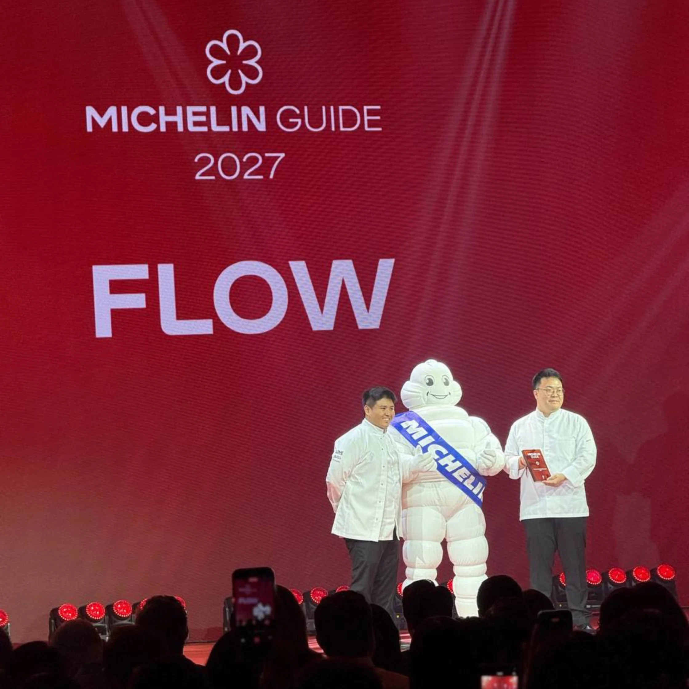 On a red Michelin Guide 2027 “FLOW” stage, two presenters stand beside the Michelin Man as audience members photograph them; the scene feels celebratory and lively.