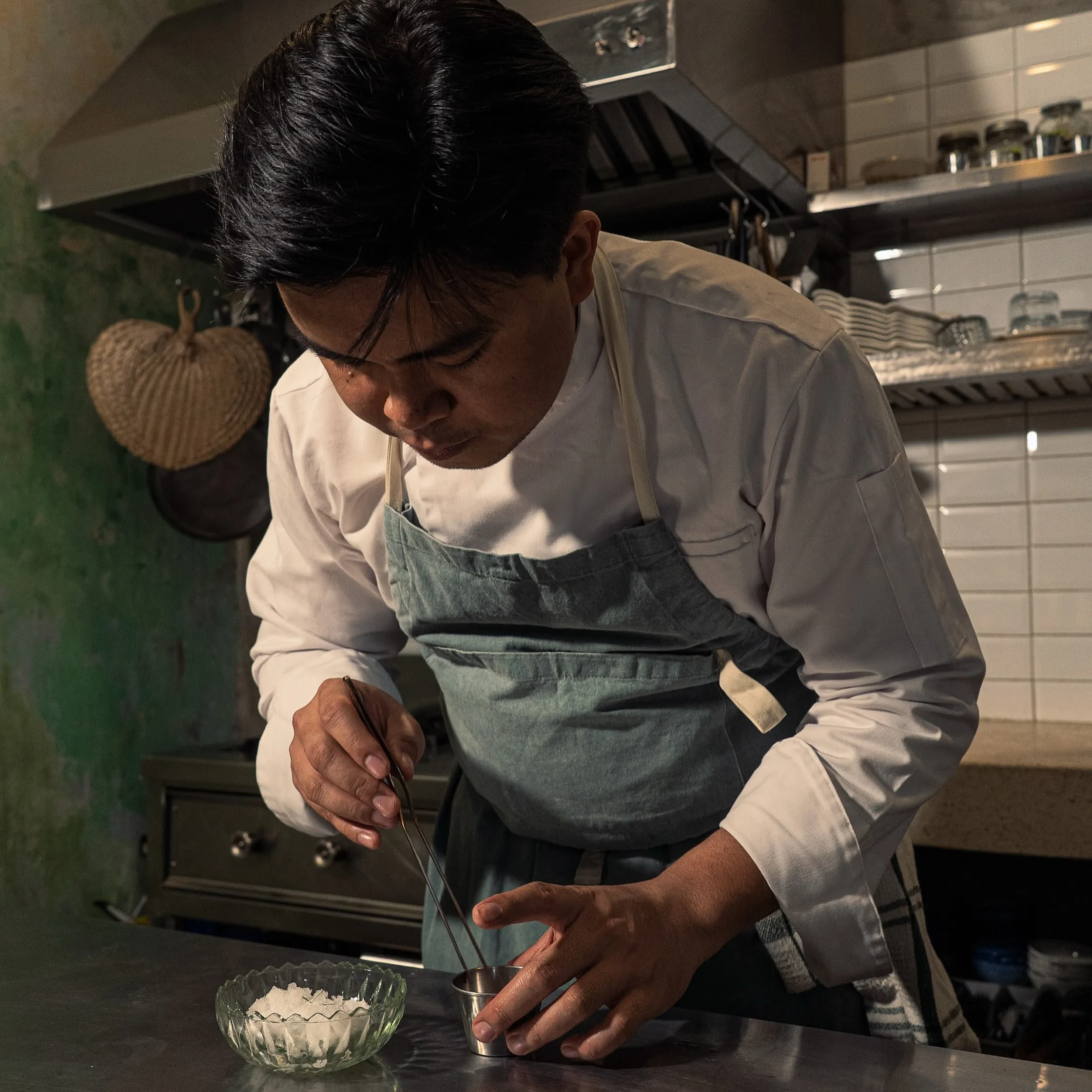 A focused chef in a white jacket and blue-gray apron carefully pours liquid into a small metal cup beside a glass bowl in a restaurant kitchen.