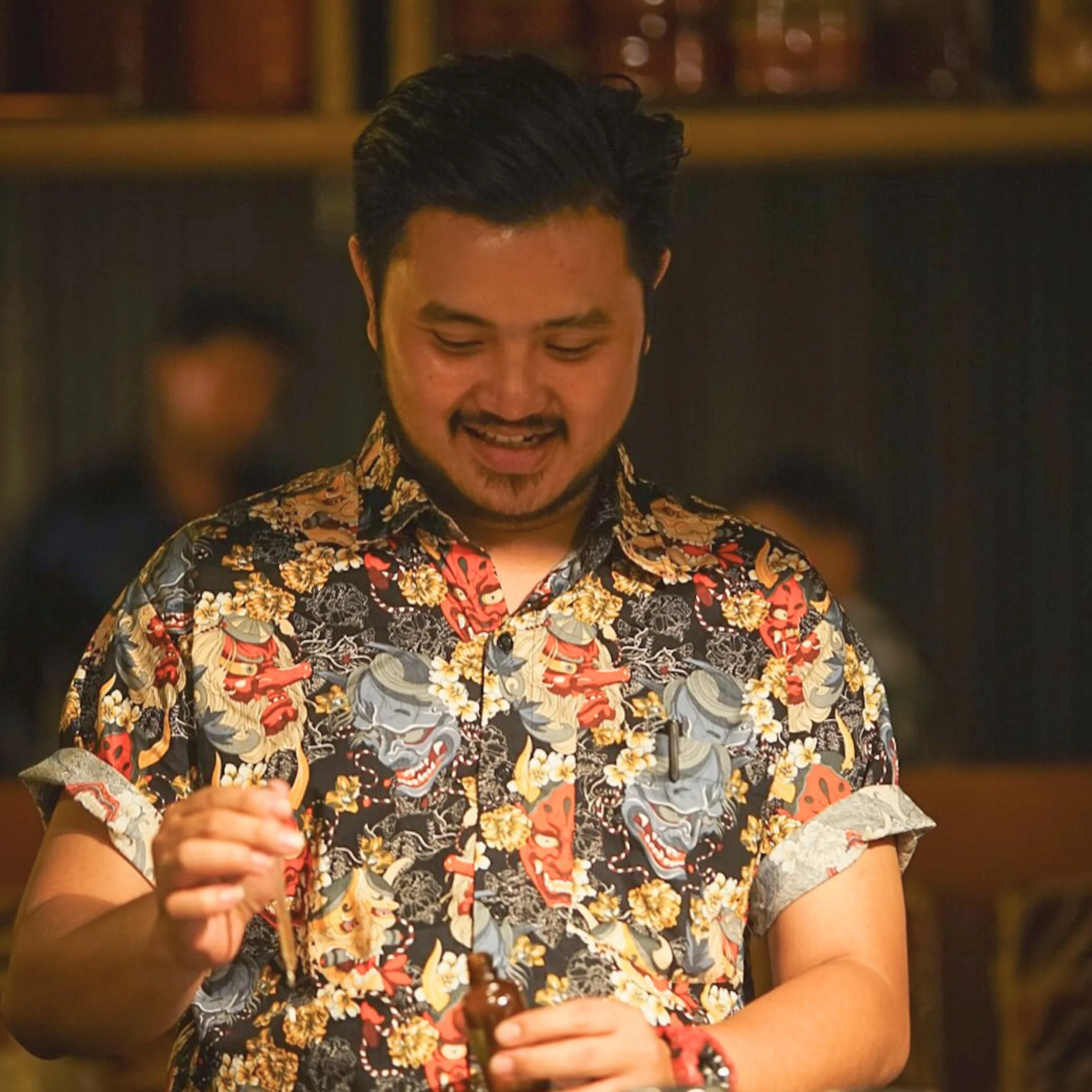 A smiling man in a colorful floral shirt looks down as he works with his hands in a warmly lit indoor setting, conveying calm focus.