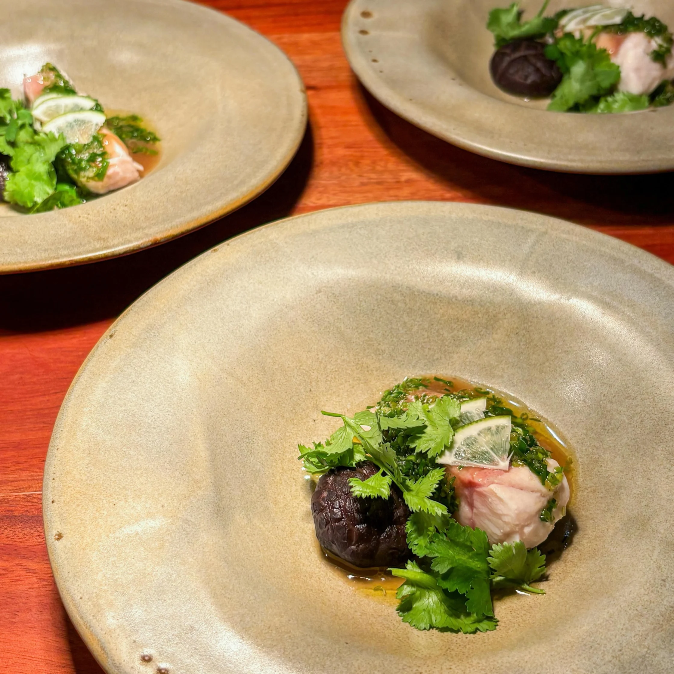 Three beige ceramic plates on a warm wooden table, each holding a small seafood dish with herbs, citrus, and dark mushrooms; elegant, intimate dining.