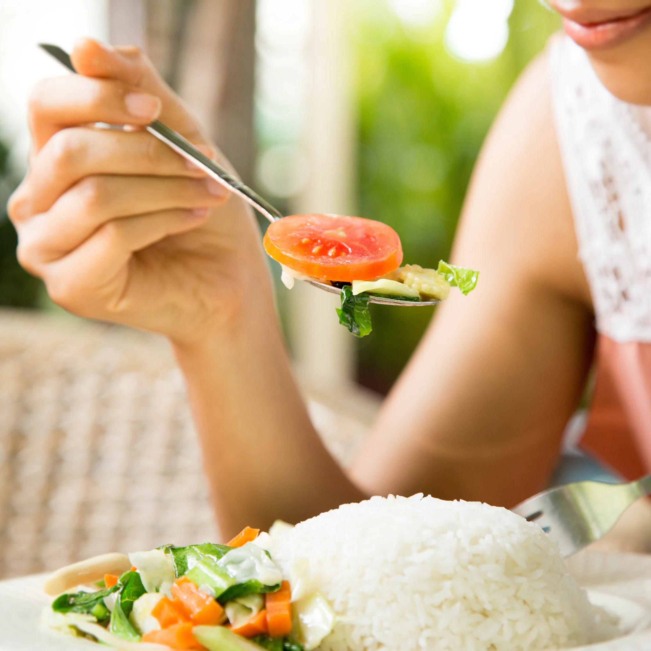 A woman lifts a forkful of tomato and greens toward her mouth beside a plate of white rice and vegetables, suggesting a fresh, relaxed meal.