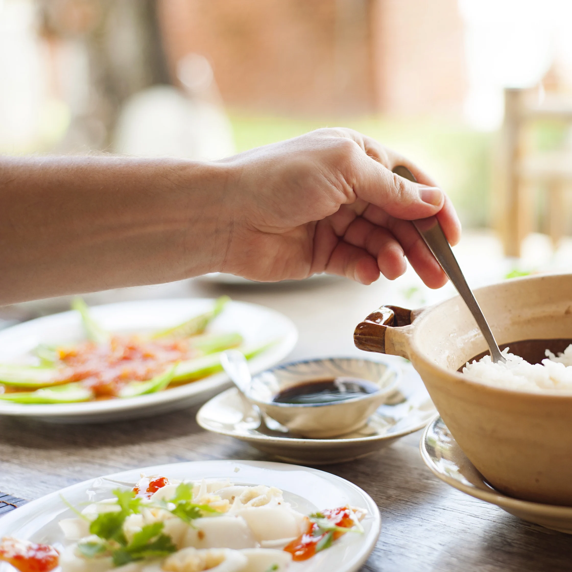 A hand uses a wooden spoon to serve white rice from a clay pot beside plated salad and dipping sauce, creating a warm, inviting meal scene.