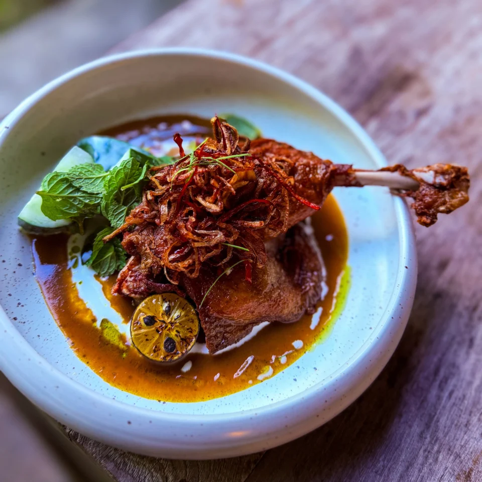 A richly spiced roasted meat shank sits in golden sauce on a pale blue plate, topped with crispy fried onions and herbs, with lime and greens; warm, rustic presentation.
