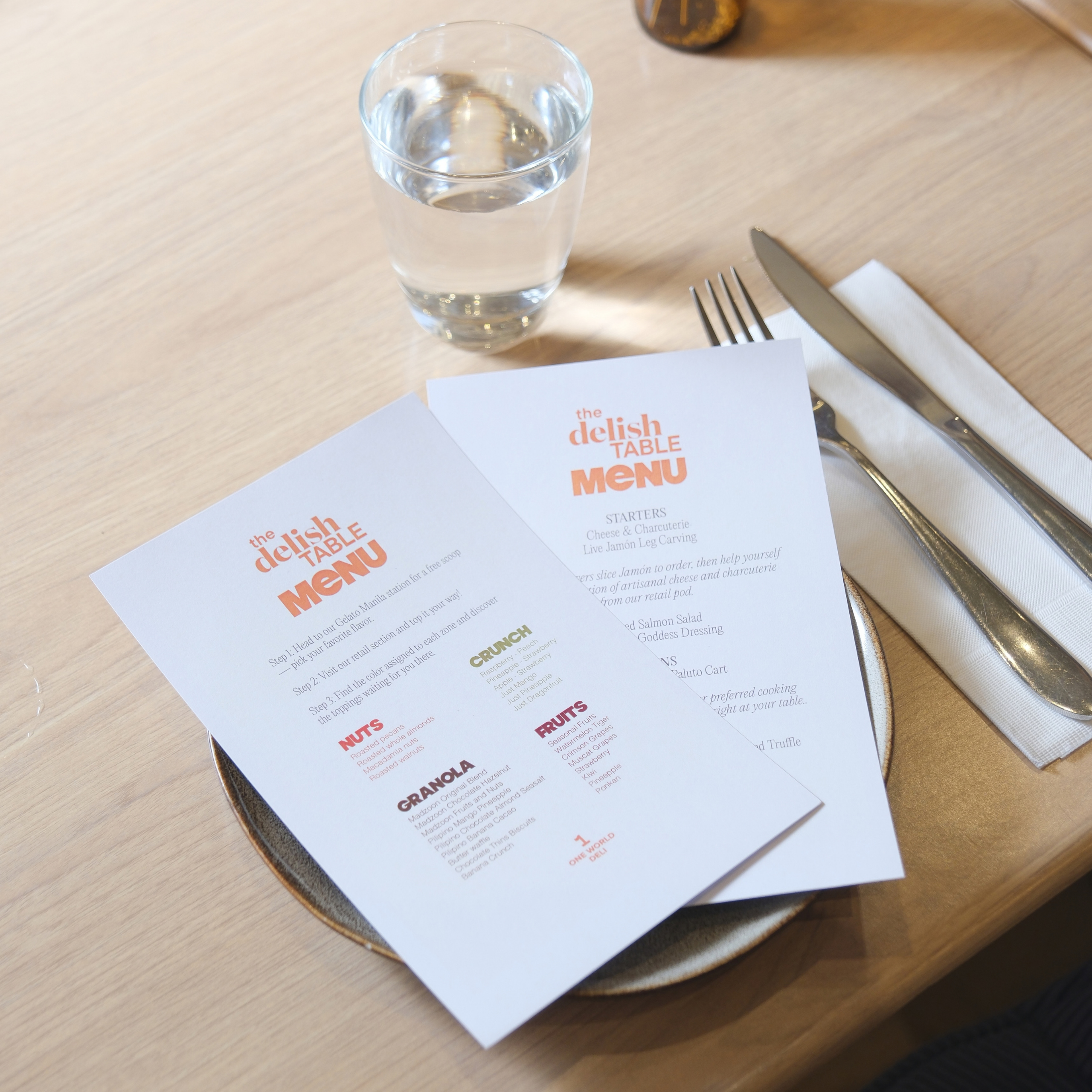 Two white “delish TABLE” menus rest on a plate beside a water glass, fork, and knife on a light wood table, creating a calm, casual dining scene.