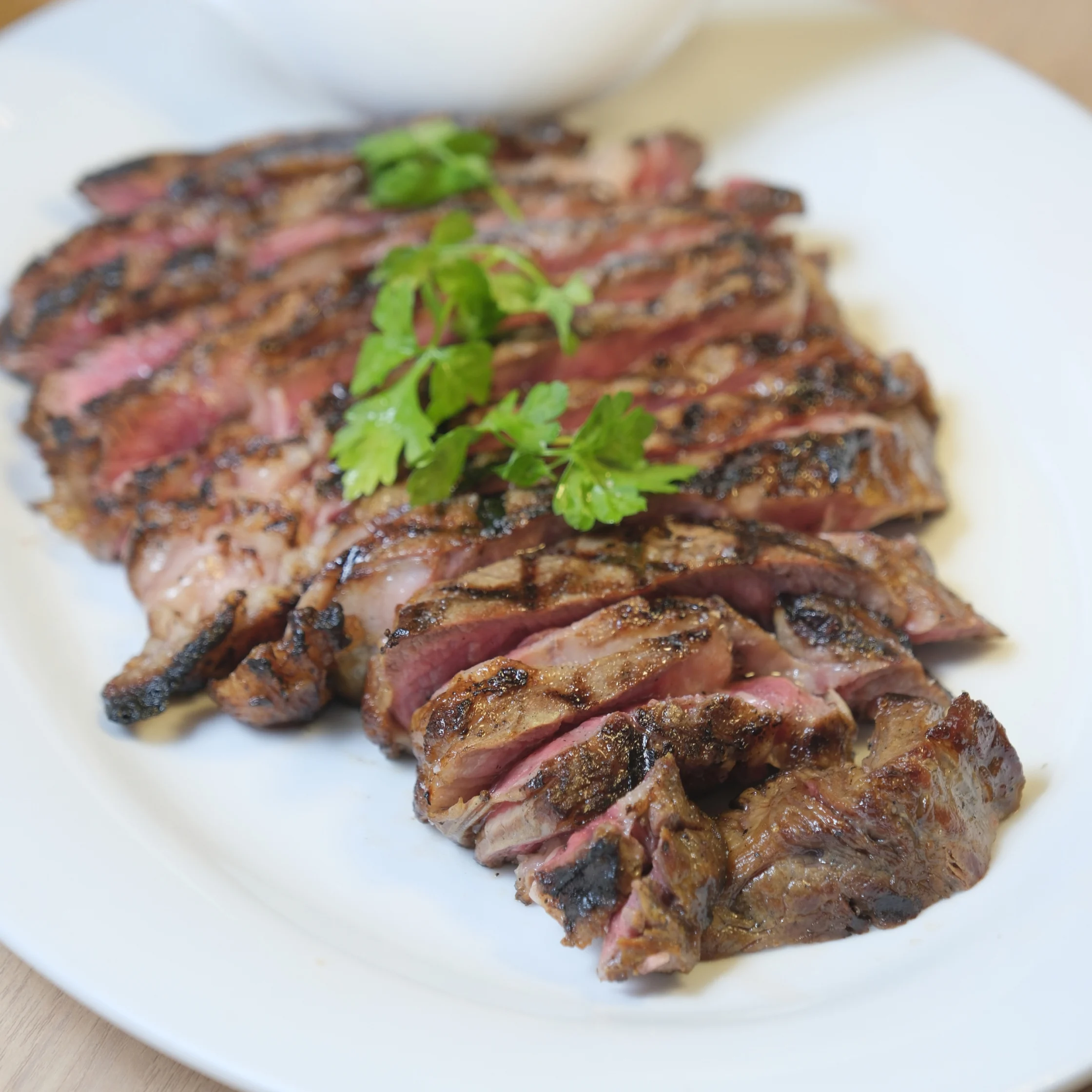 Sliced grilled steak with charred edges and a pink center, arranged on a white plate and topped with fresh parsley; a hearty, appetizing dish.