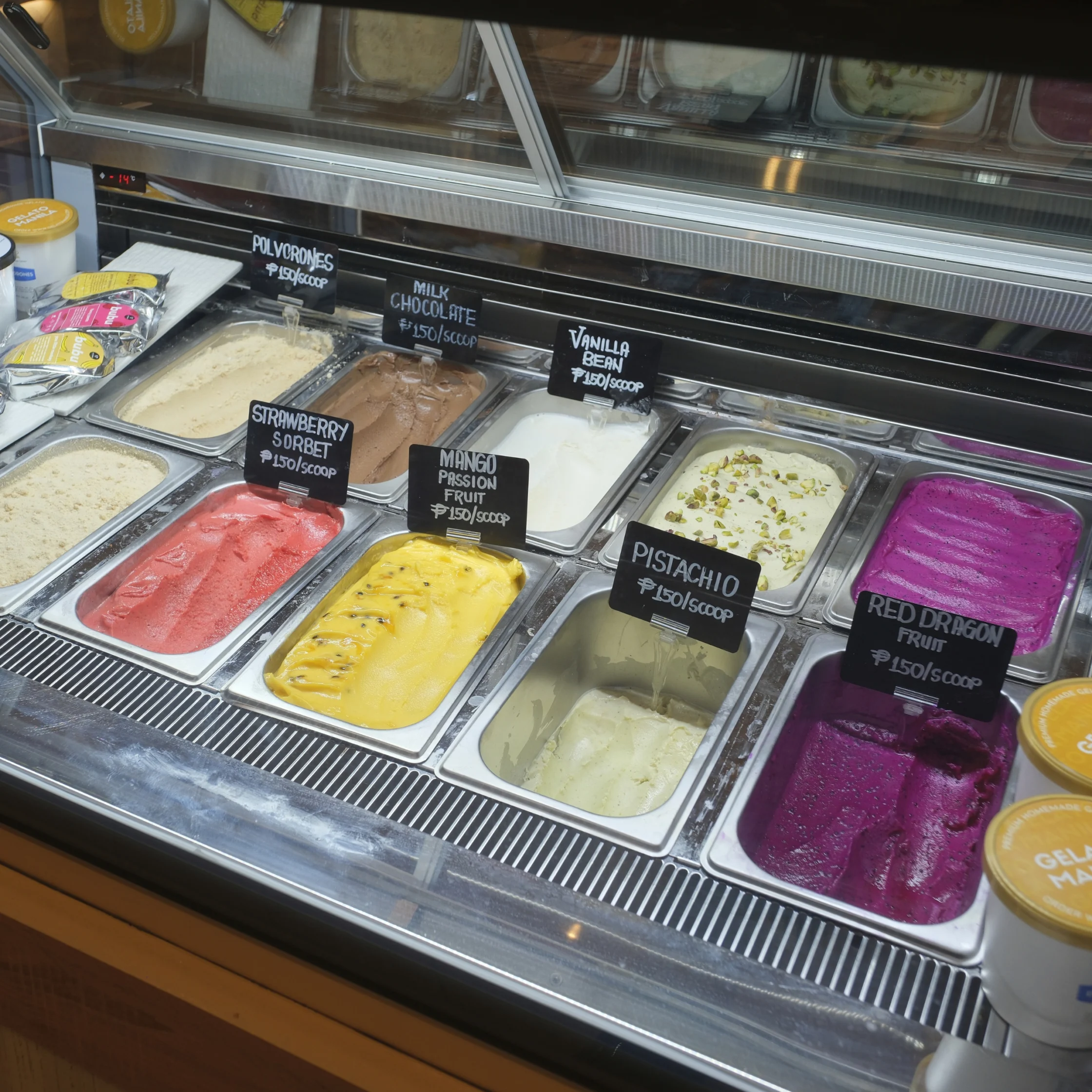 A gelato display case holds colorful tubs of strawberry sorbet, mango passion fruit, pistachio, red dragon fruit, vanilla bean, and chocolate; inviting dessert counter.
