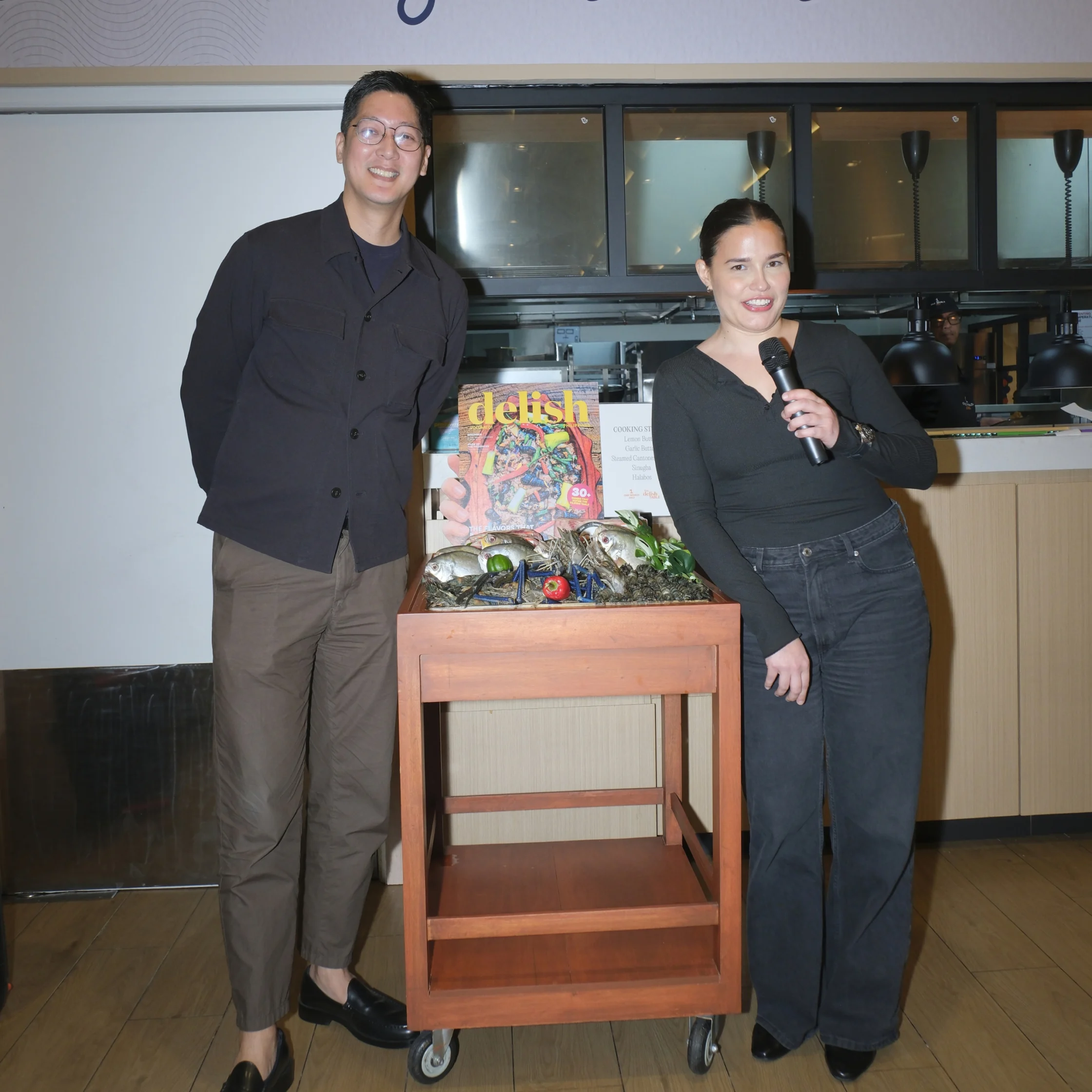 Two smiling presenters pose beside a seafood display and “Delish” magazine in a restaurant kitchen; one holds a microphone, creating a cheerful event atmosphere.