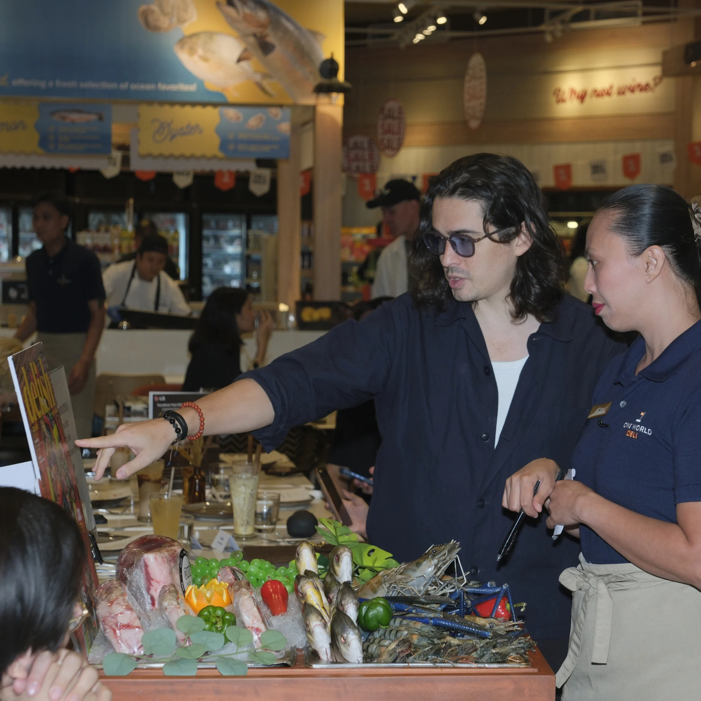 A man in sunglasses points out seafood at a busy market stall while a female worker listens; fish, shellfish, and colorful produce fill the counter.