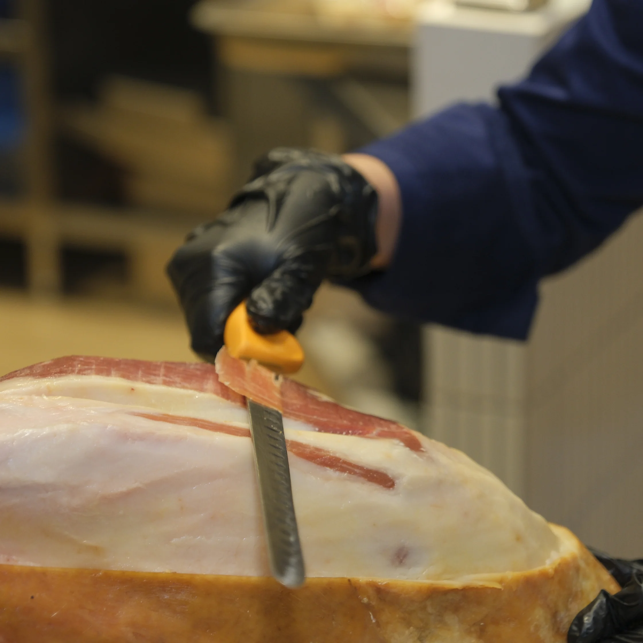 A black-gloved hand uses an orange-handled knife to score a large cured ham, revealing pale fat and red meat in a butcher’s workspace. The scene feels focused and practical.