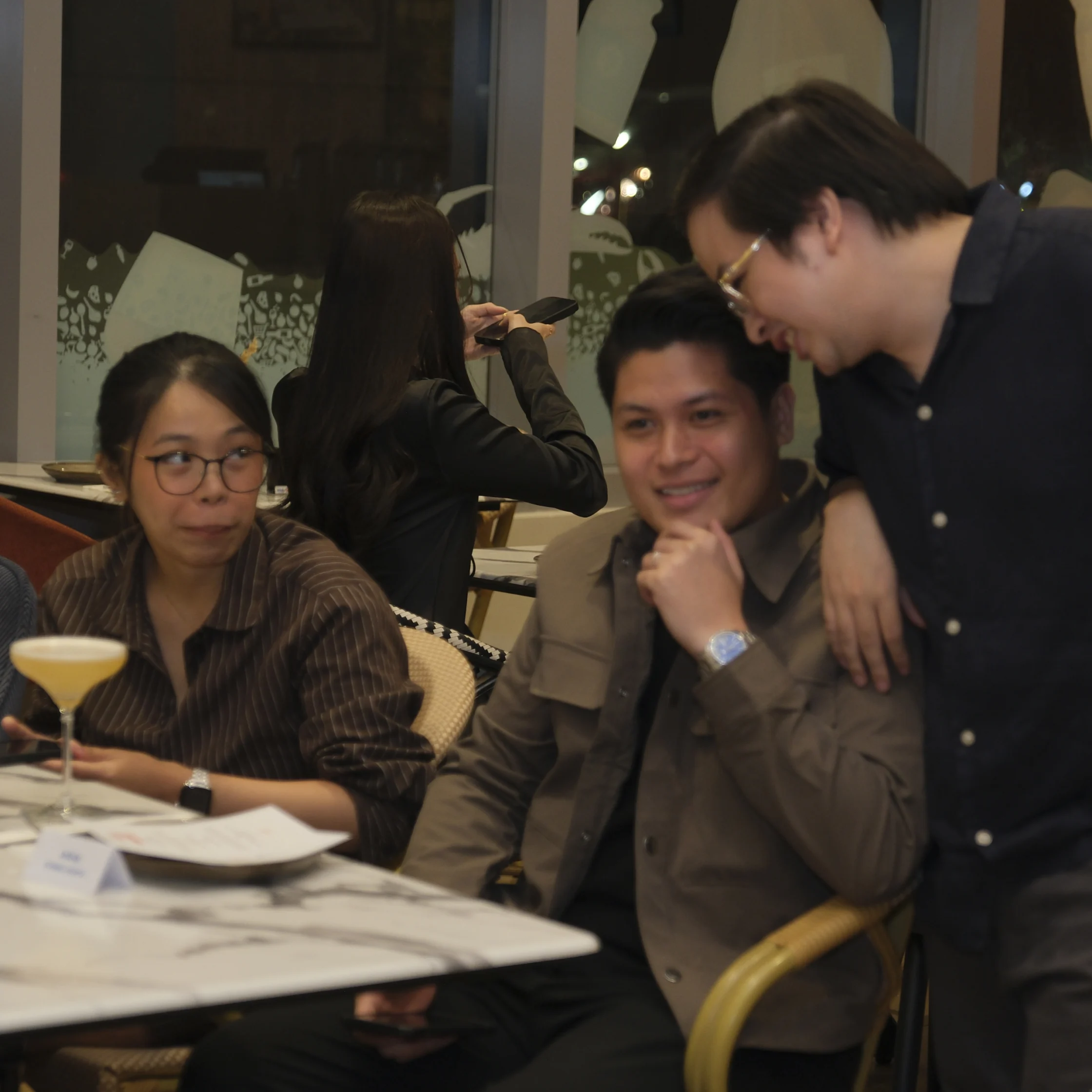 At a restaurant table, a woman looks aside while a seated man smiles and a standing man leans in to talk; the scene feels relaxed and social.