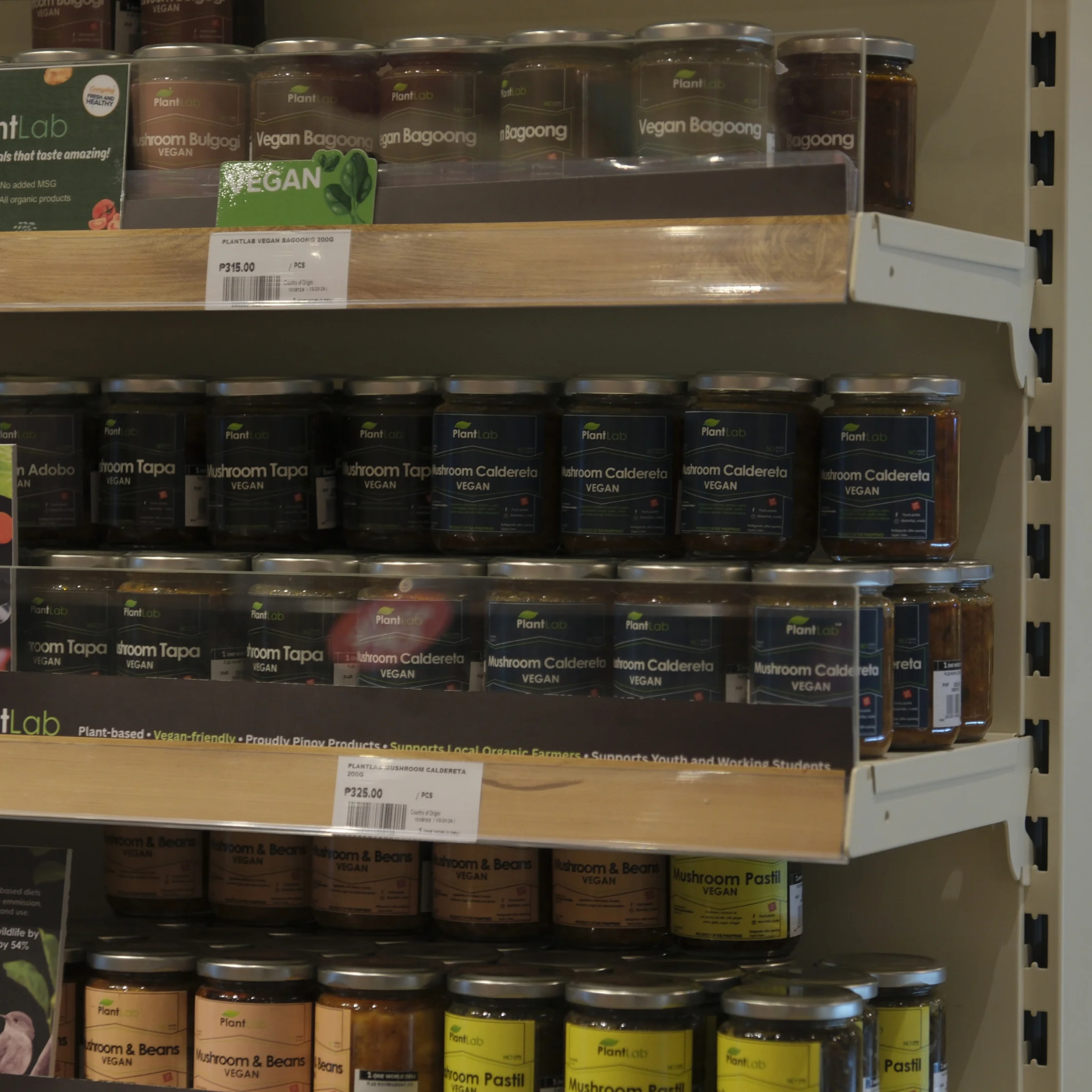 Rows of labeled jars of vegan condiments and spreads fill grocery shelves, including vegan bagoong, tapenade, and mushroom pastes; the scene feels practical and product-focused.
