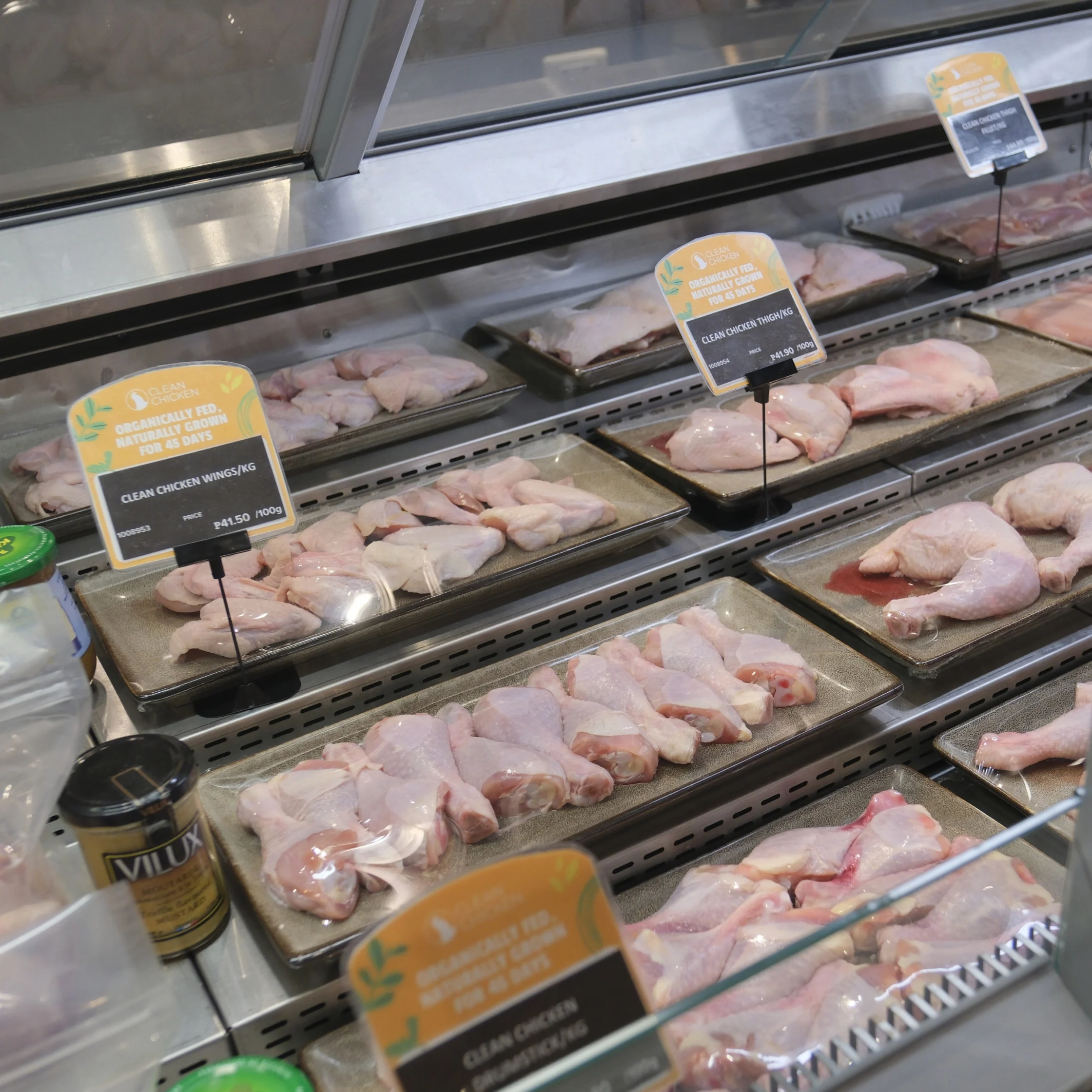 Raw chicken portions on metal trays in a refrigerated butcher display, with price labels and glass panels; a clean, orderly grocery counter.