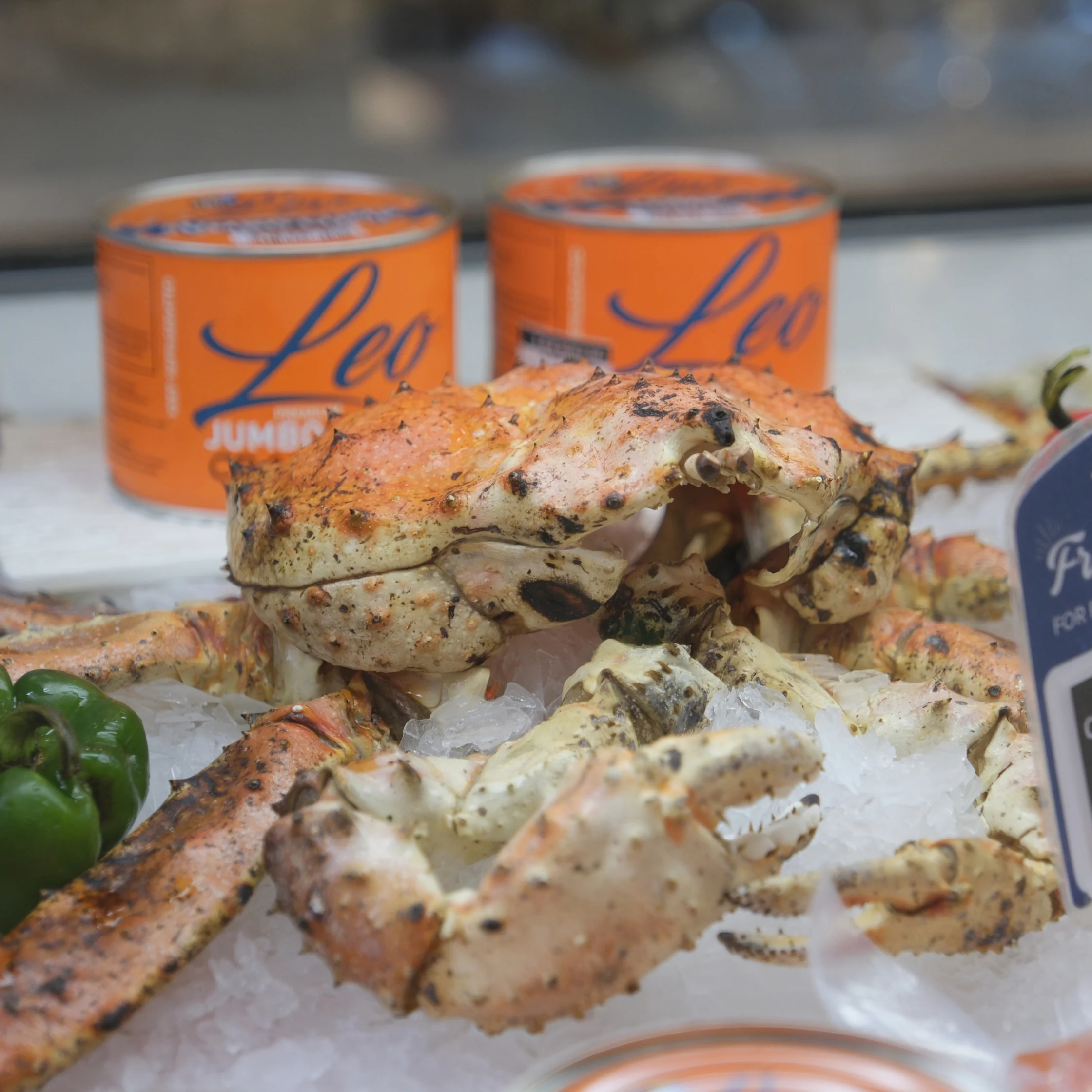 Grilled crab legs rest on crushed ice beside green pepper, with two orange Leo cans behind them; the market display feels fresh and appetizing.