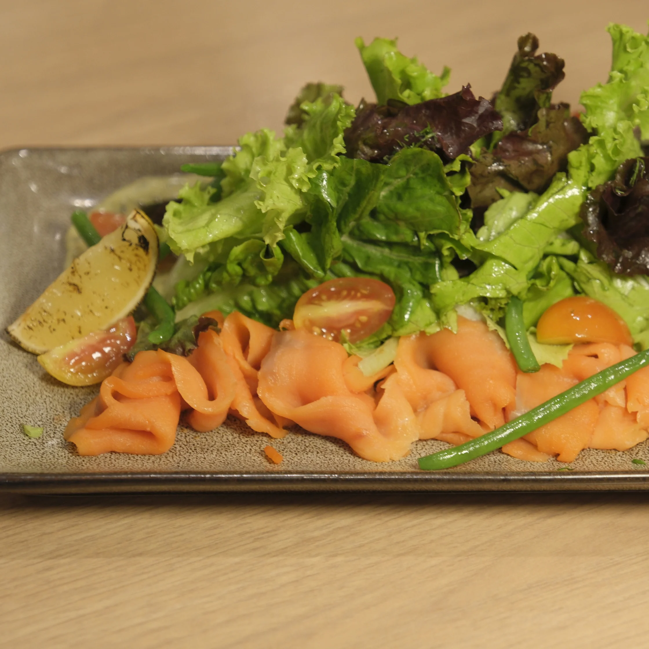 A colorful salad on a rectangular ceramic plate features folded smoked salmon, leafy greens, cherry tomatoes, lemon wedges, and green beans, arranged fresh and inviting.