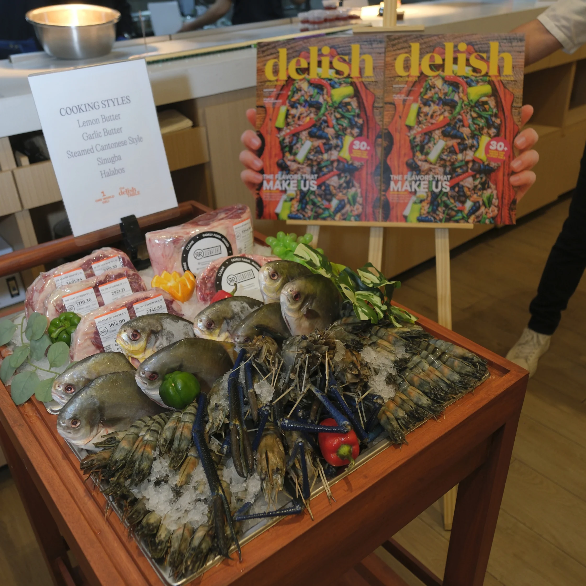 A seafood market display of fish, prawns, and lobster on ice with lemons and herbs, beside a person holding two Delish magazines; inviting, colorful food photography.