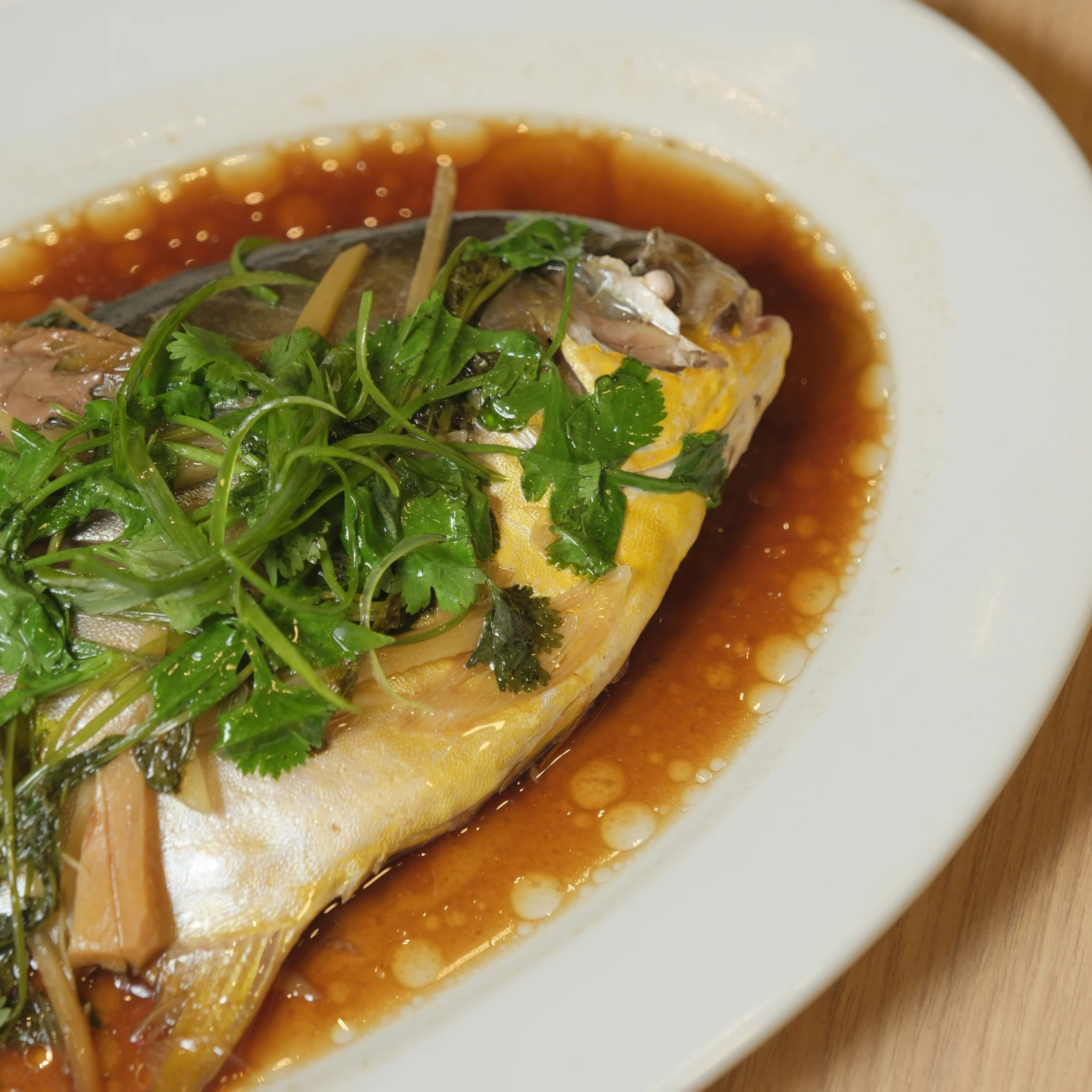 A whole steamed fish topped with fresh cilantro and scallions rests in glossy brown sauce on a white oval plate, evoking a savory Asian meal.