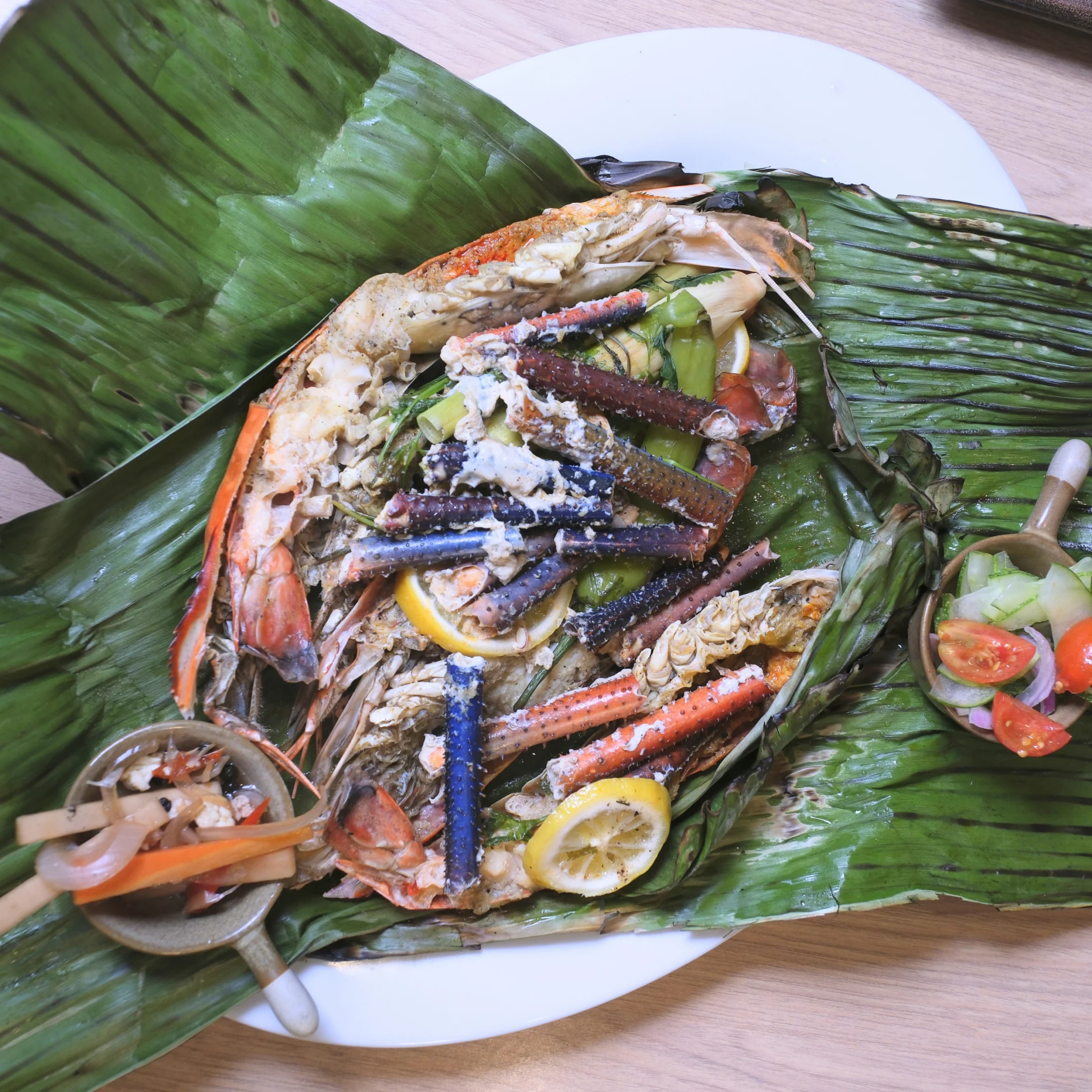A banana-leaf-lined platter piled with grilled seafood, including shrimp, fish, crab, and lemon slices, with dipping sauce and fresh salad; abundant and festive.