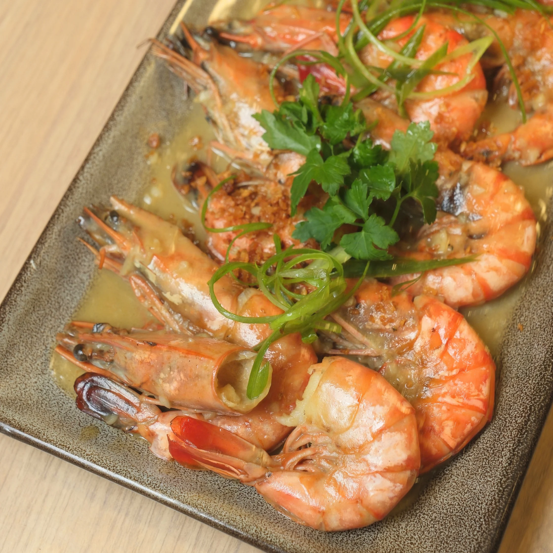 A dark ceramic platter holds orange prawns in savory sauce, topped with fresh parsley and curled green onion; the dish looks warm and appetizing.