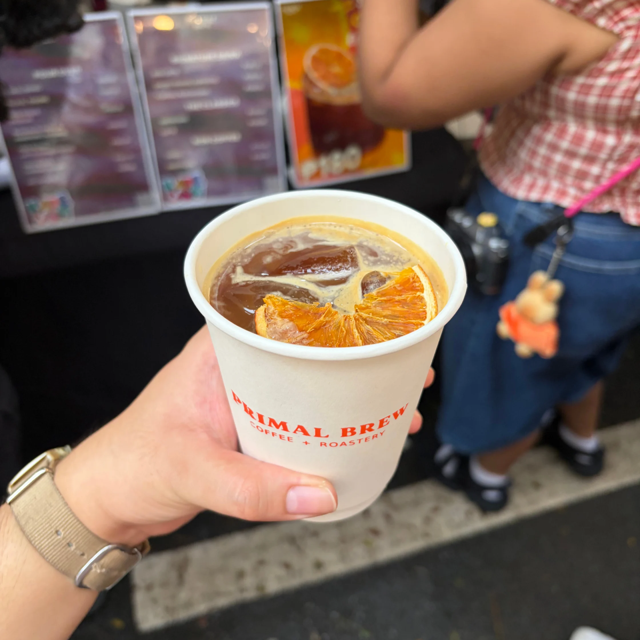 Hand holding a white paper cup of iced coffee garnished with orange slices, in front of a café menu and a customer; the scene feels casual and inviting.