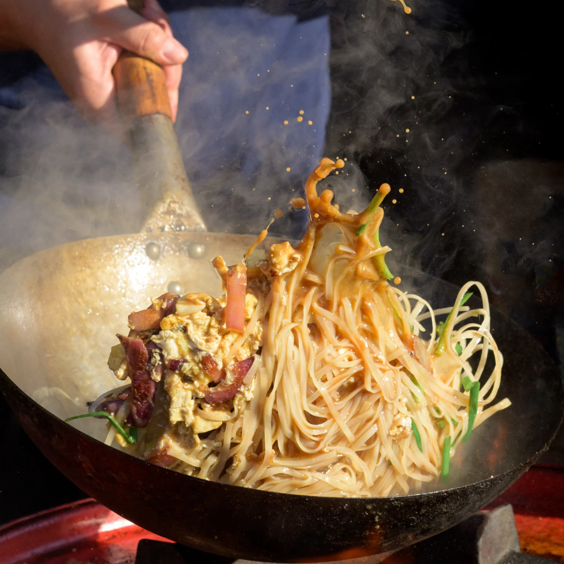 A cook tosses steaming noodles, vegetables, and egg in a hot wok, sending sauce and ingredients into the air; the scene feels energetic and appetizing.