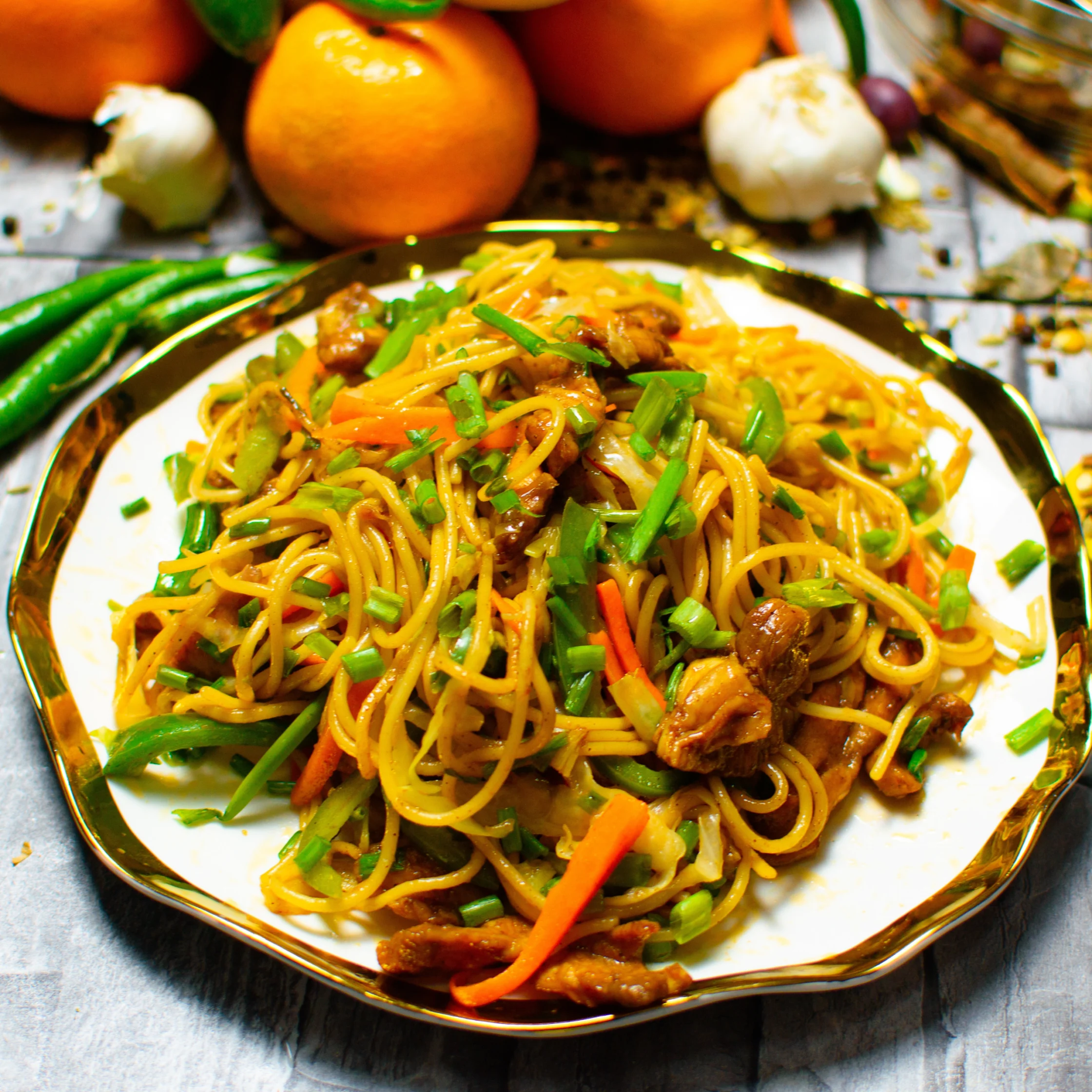 A generous plate of golden stir-fried noodles with vegetables and browned meat, garnished with herbs; oranges, garlic, and green chilies frame the appetizing dish.