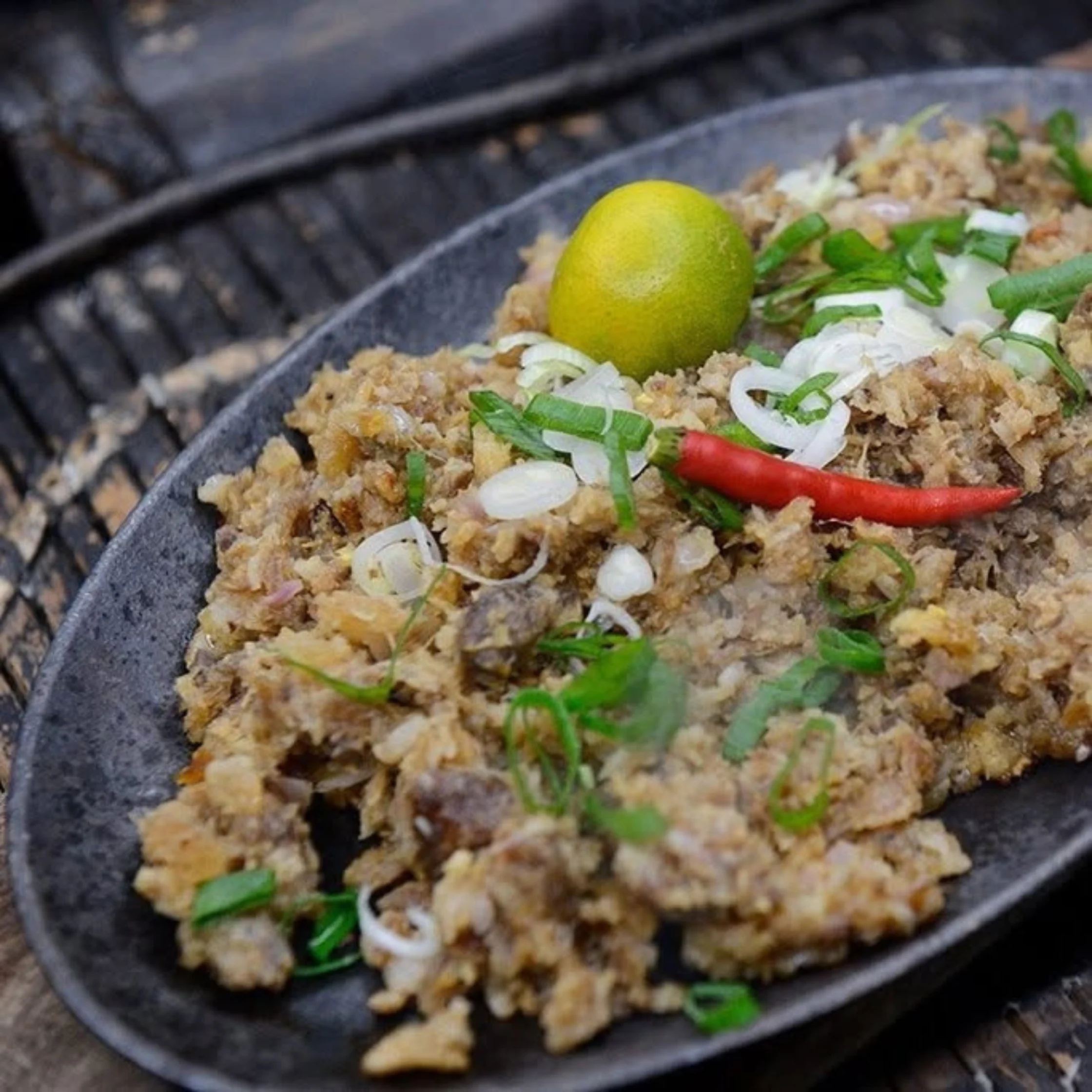 A rustic black platter holds chopped, browned meat topped with scallions, sliced onion, a red chili, and lime; the grilled dish looks savory and rustic.