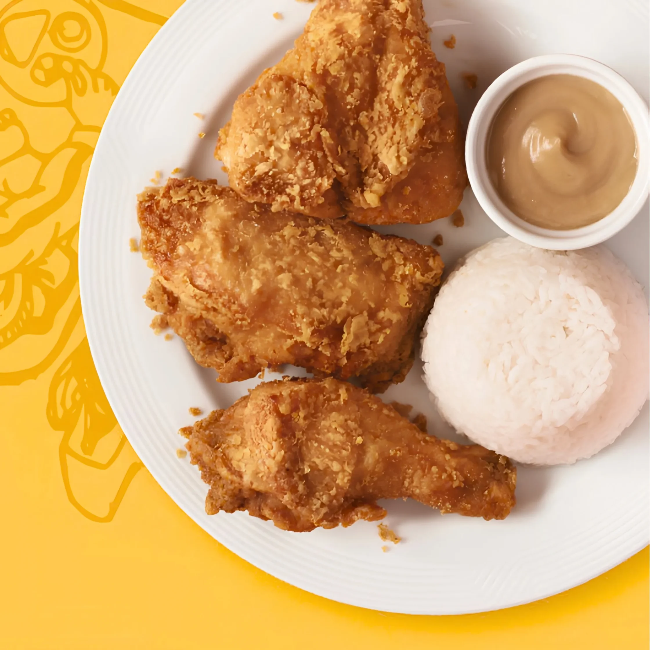 Golden, crispy fried chicken pieces, a molded mound of white rice, and creamy dipping sauce on a white plate over a bright yellow patterned background.