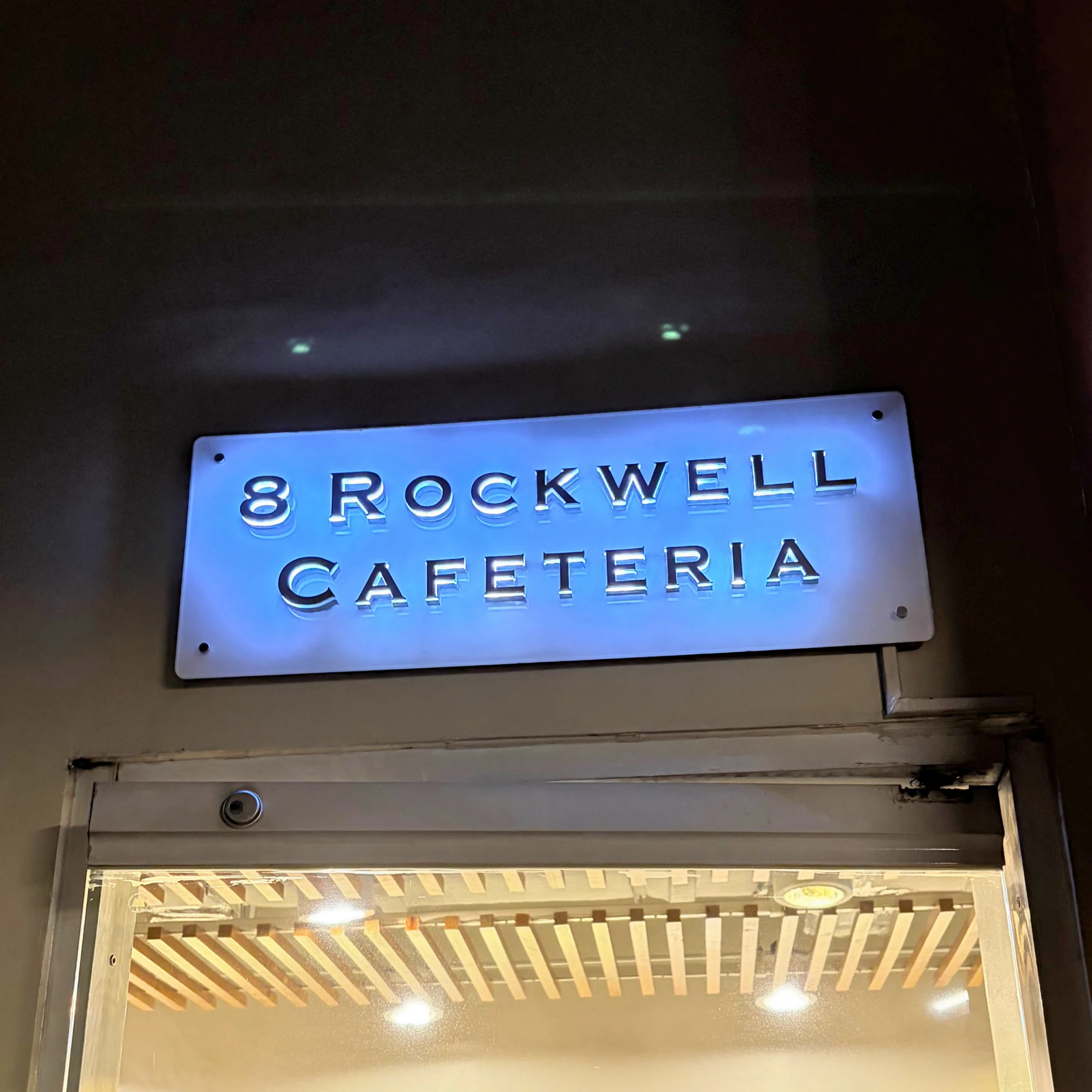 A glowing blue “8 Rockwell Cafeteria” sign hangs above a warmly lit entrance with a slatted ceiling, creating a quiet nighttime atmosphere.