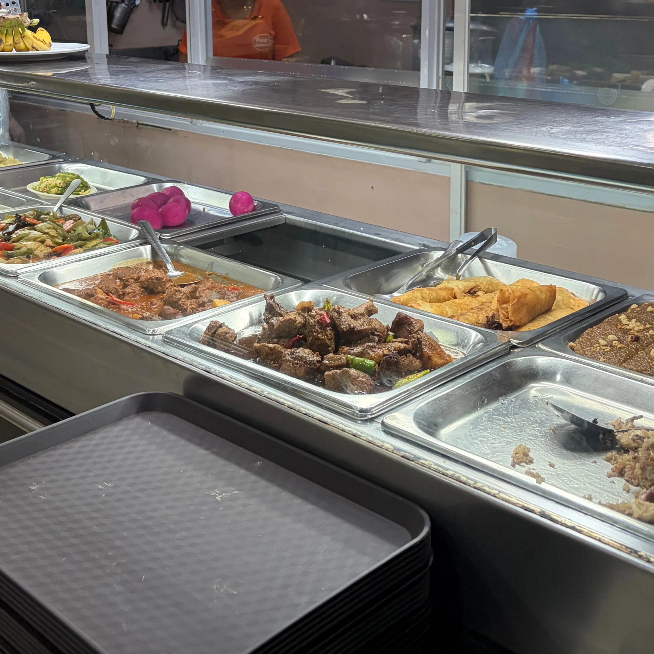 A cafeteria buffet displays trays of stewed meat, fried pastries, vegetables, and rice behind a glass counter, with serving spoons and stacked trays.