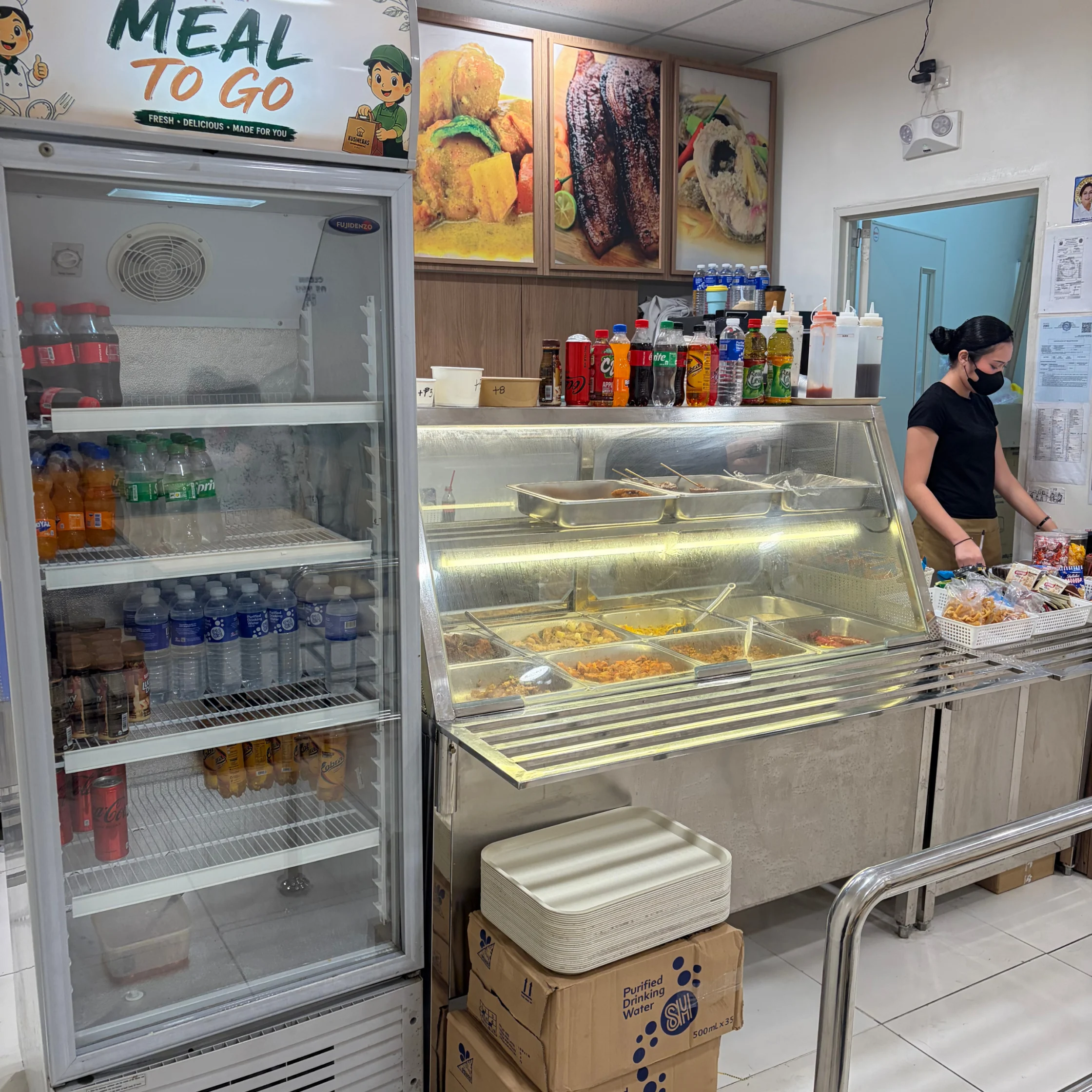 A small deli counter displays chilled drinks and hot prepared foods behind glass, with sandwich photos above and a masked worker serving customers.