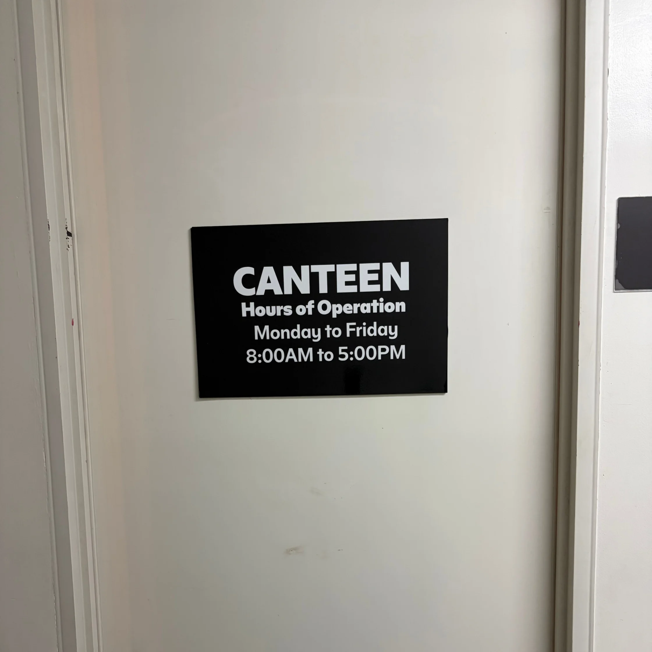 A black sign on a white door reads “CANTEEN” and lists weekday hours, 8:00 AM–5:00 PM; the plain setting feels quiet and practical.