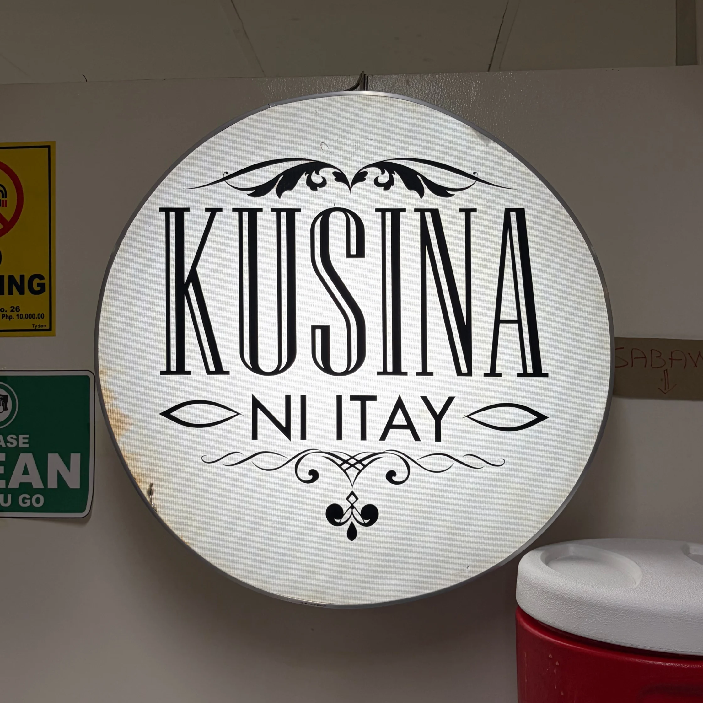 A glowing round wall sign reads “Kusina ni Itay” in ornate black lettering with decorative flourishes, creating a warm, welcoming restaurant feel.