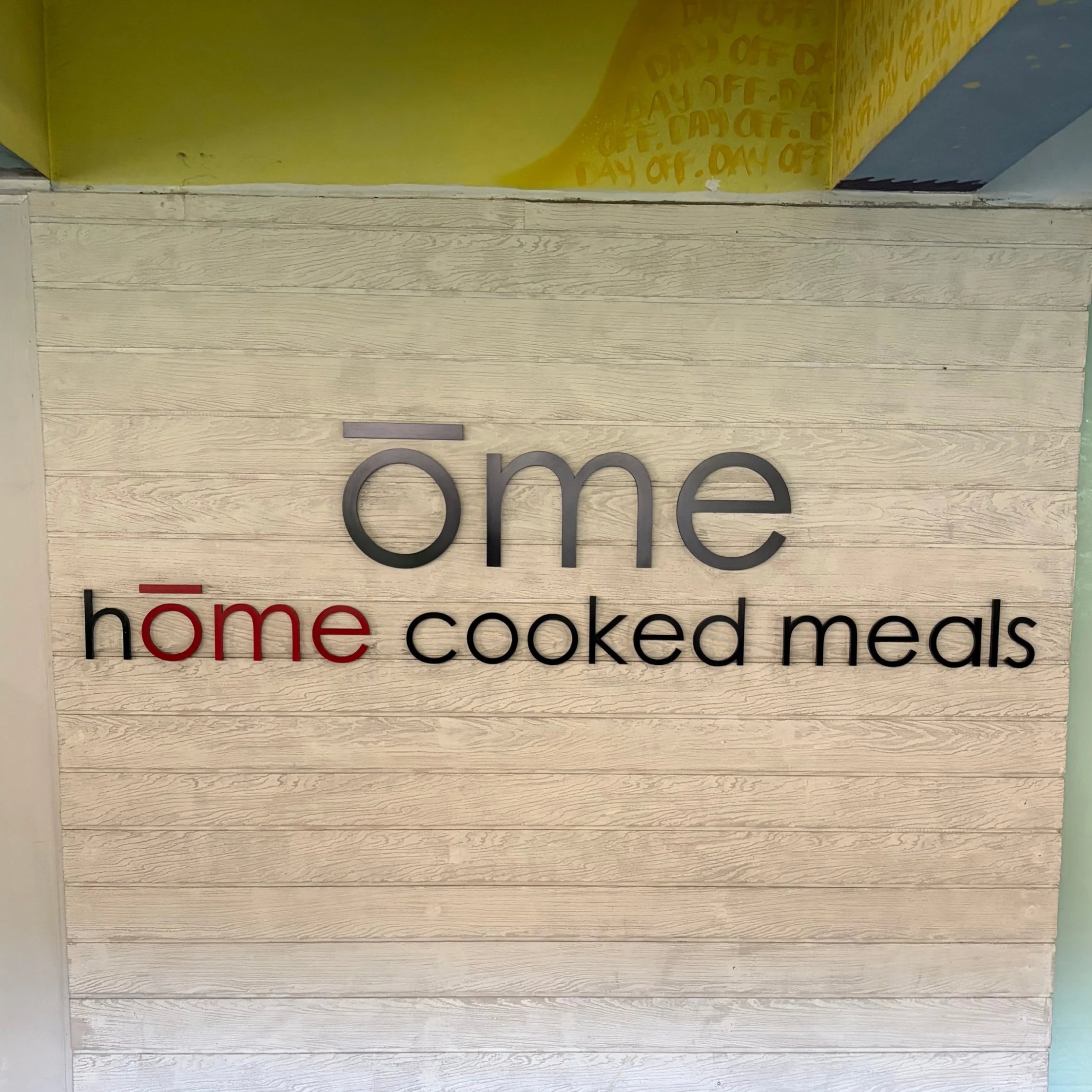 Restaurant sign reading “ōme home cooked meals” on pale wood paneling, with gray and red lettering; the warm, simple design feels welcoming and homey.