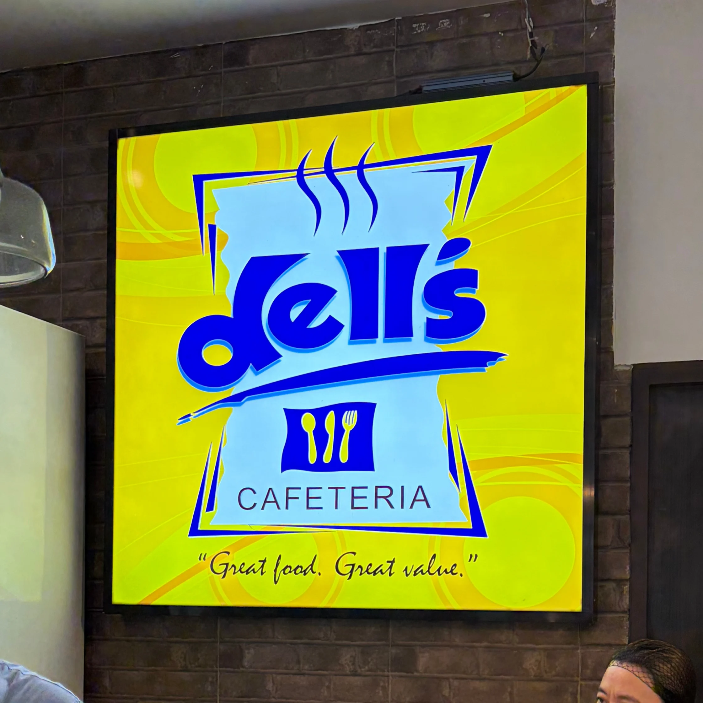 Bright yellow “Dell’s Cafeteria” sign on a dark brick wall, with a blue steaming-dish logo and the slogan “Great food. Great value.” cheerful and welcoming.