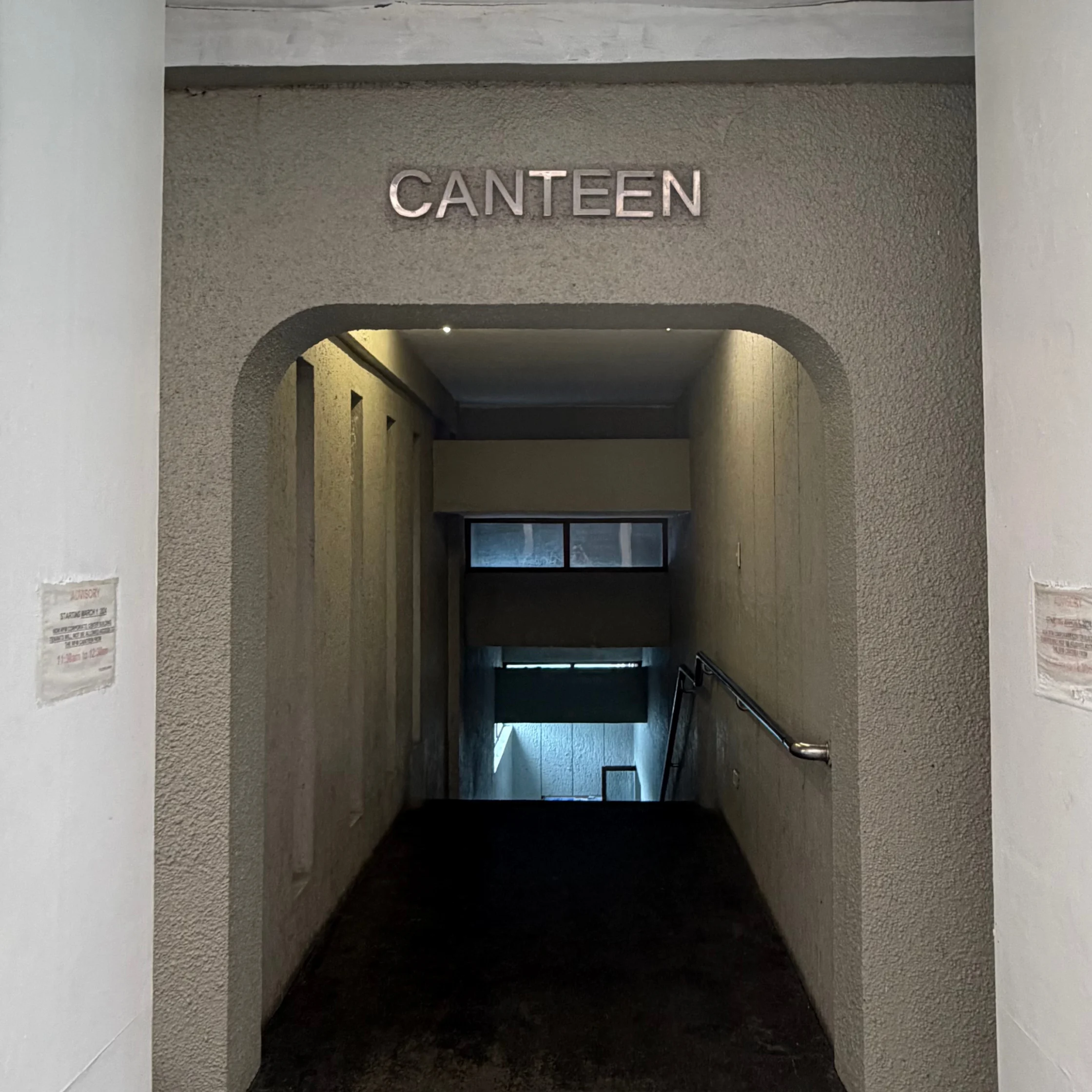 A dim concrete passage descends beneath an arched entrance marked “CANTEEN,” with a metal handrail and faint window light, creating a quiet, austere mood.
