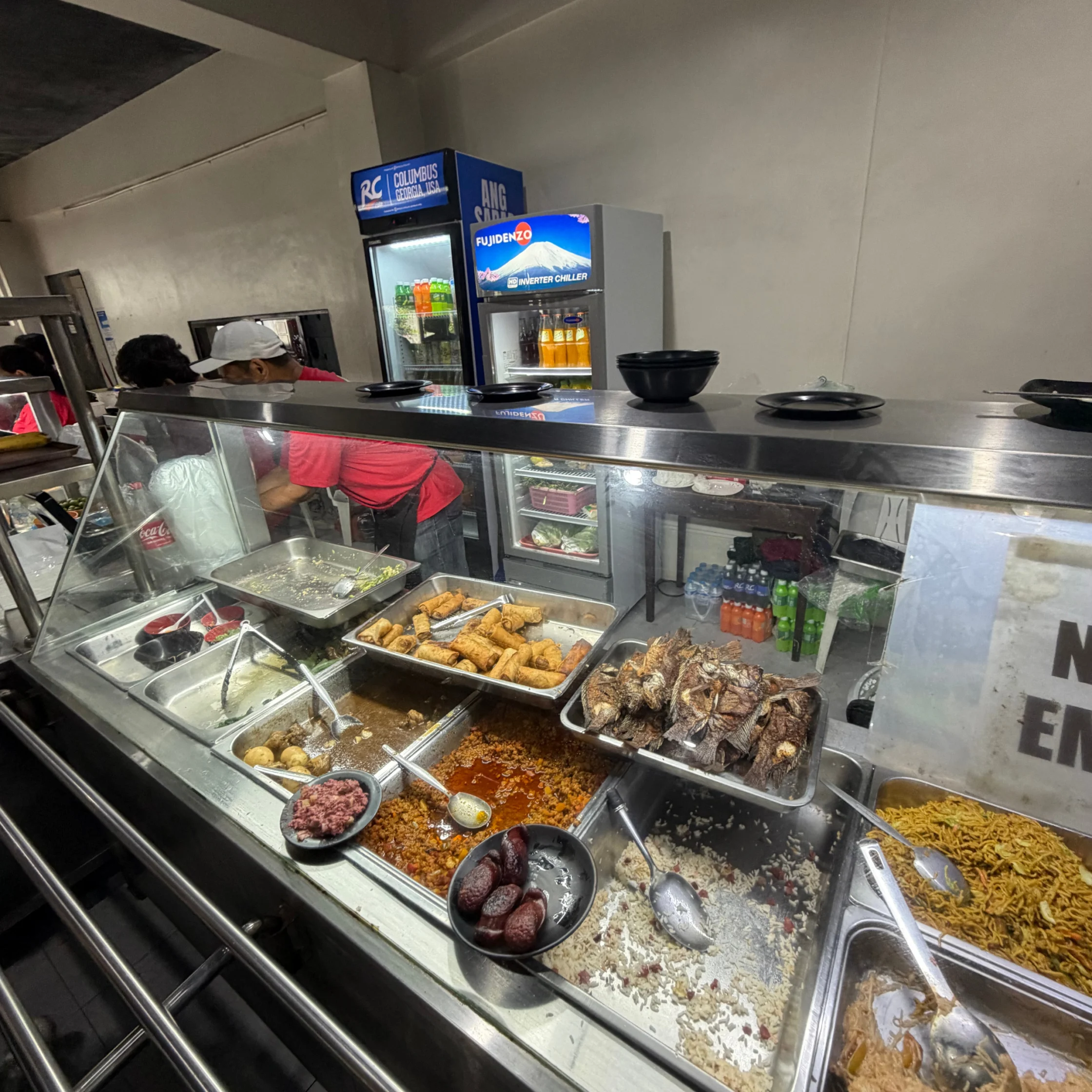 Glass-fronted restaurant buffet displays trays of rice, stews, fried snacks, grilled meat, and sausages as a worker serves food; casual, busy atmosphere.