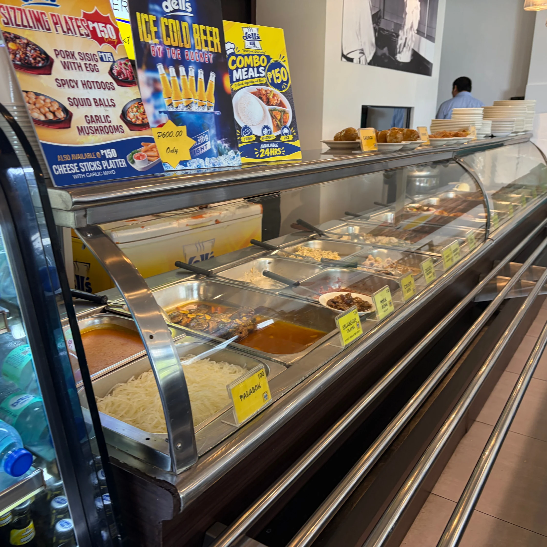 A brightly lit deli counter displays trays of prepared food beneath colorful sandwich and meal deal signs, with a worker and stacks of plates in the background.