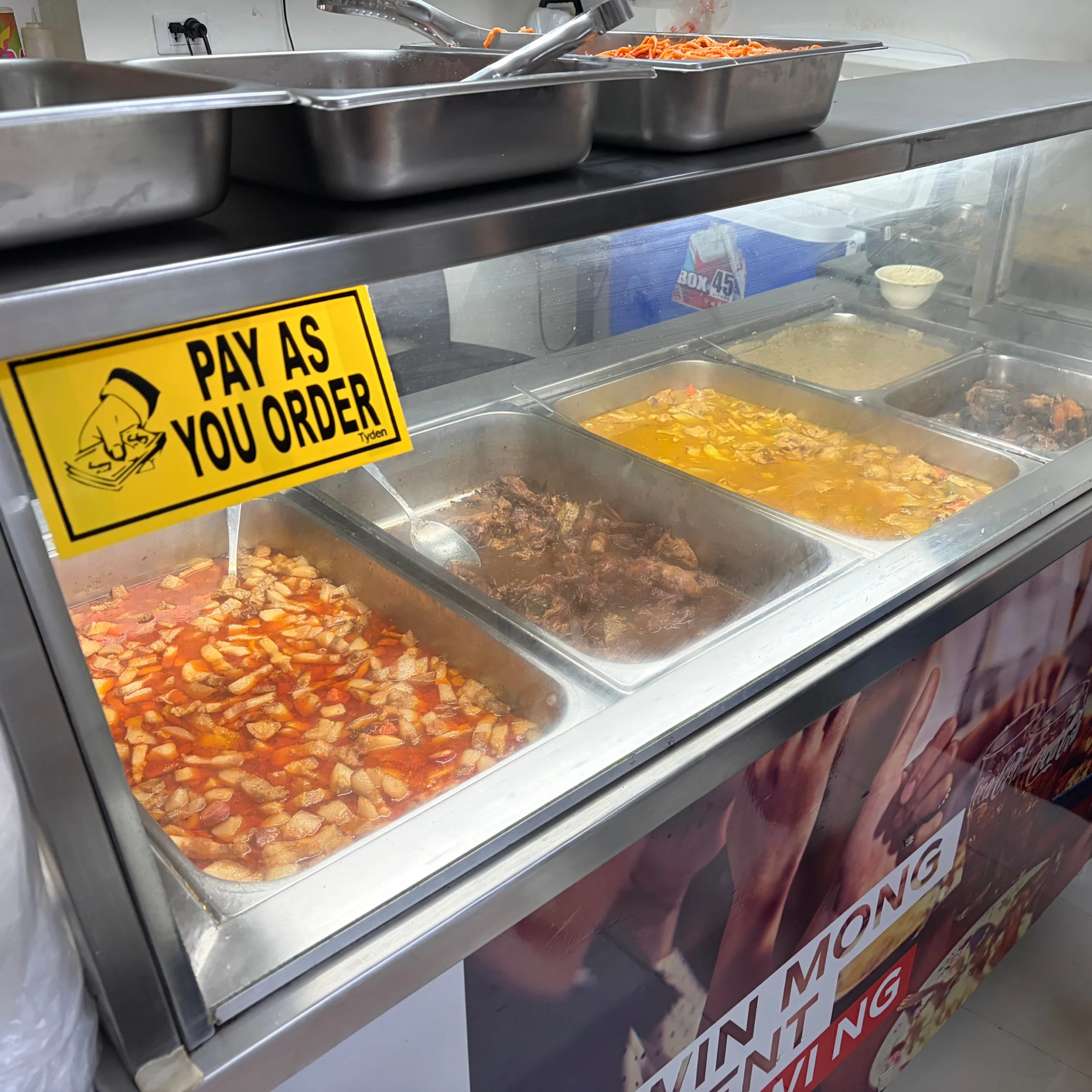 A stainless-steel food counter displays trays of saucy dishes behind glass, with a bright yellow “Pay as you order” sign; the casual tone suggests a busy canteen.